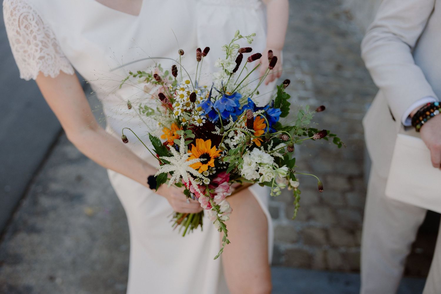 Bride holding a vibrant wildflower wedding bouquet with blue, yellow, and white blooms in an outdoor setting.