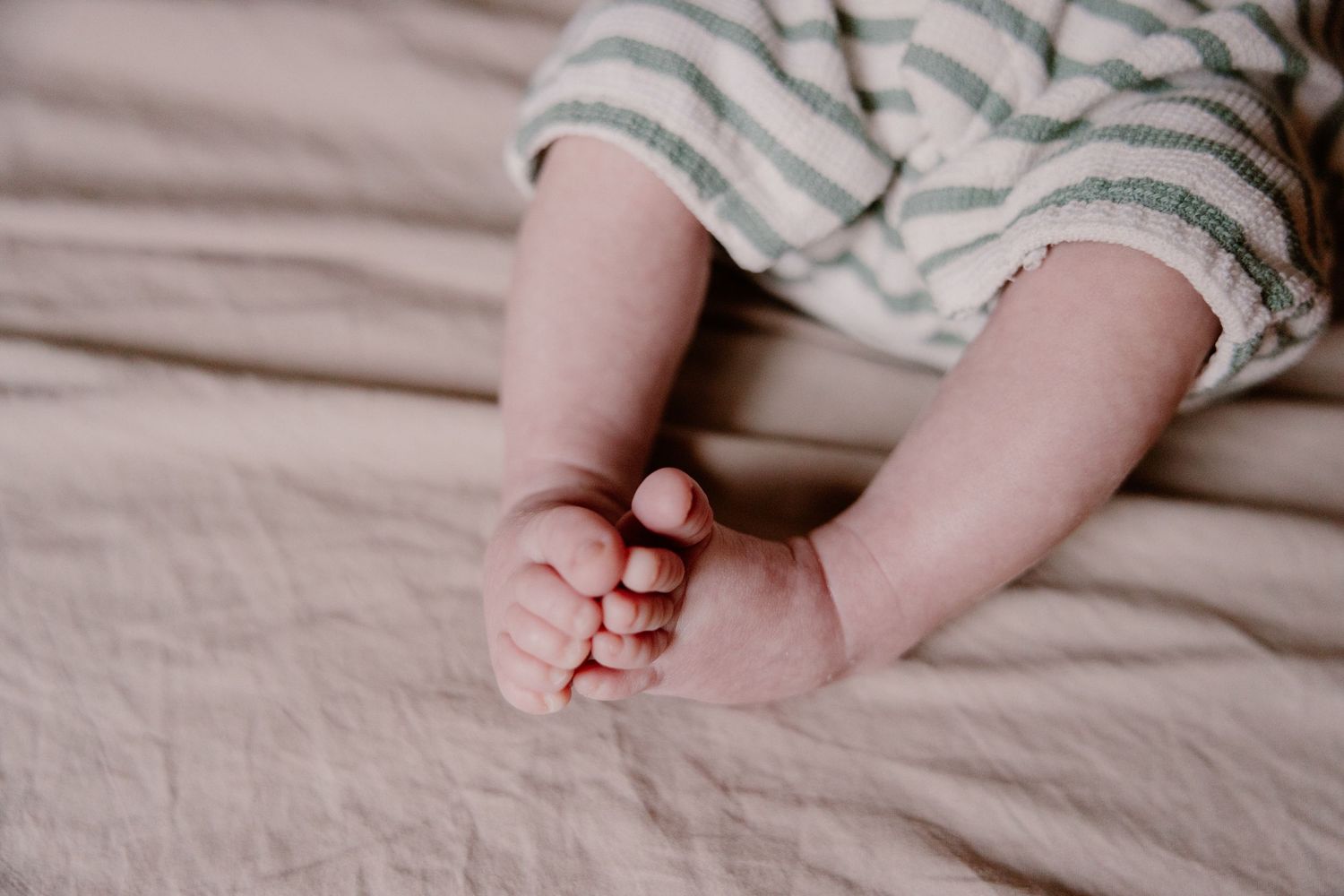 Close-up of tiny baby feet resting on a soft beige linen blanket, wearing striped pants.