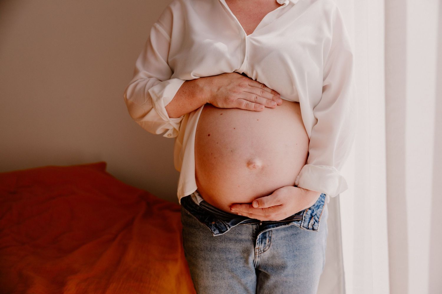 Pregnant woman in white shirt and jeans cradling bare baby bump, standing near window in cozy bedroom.