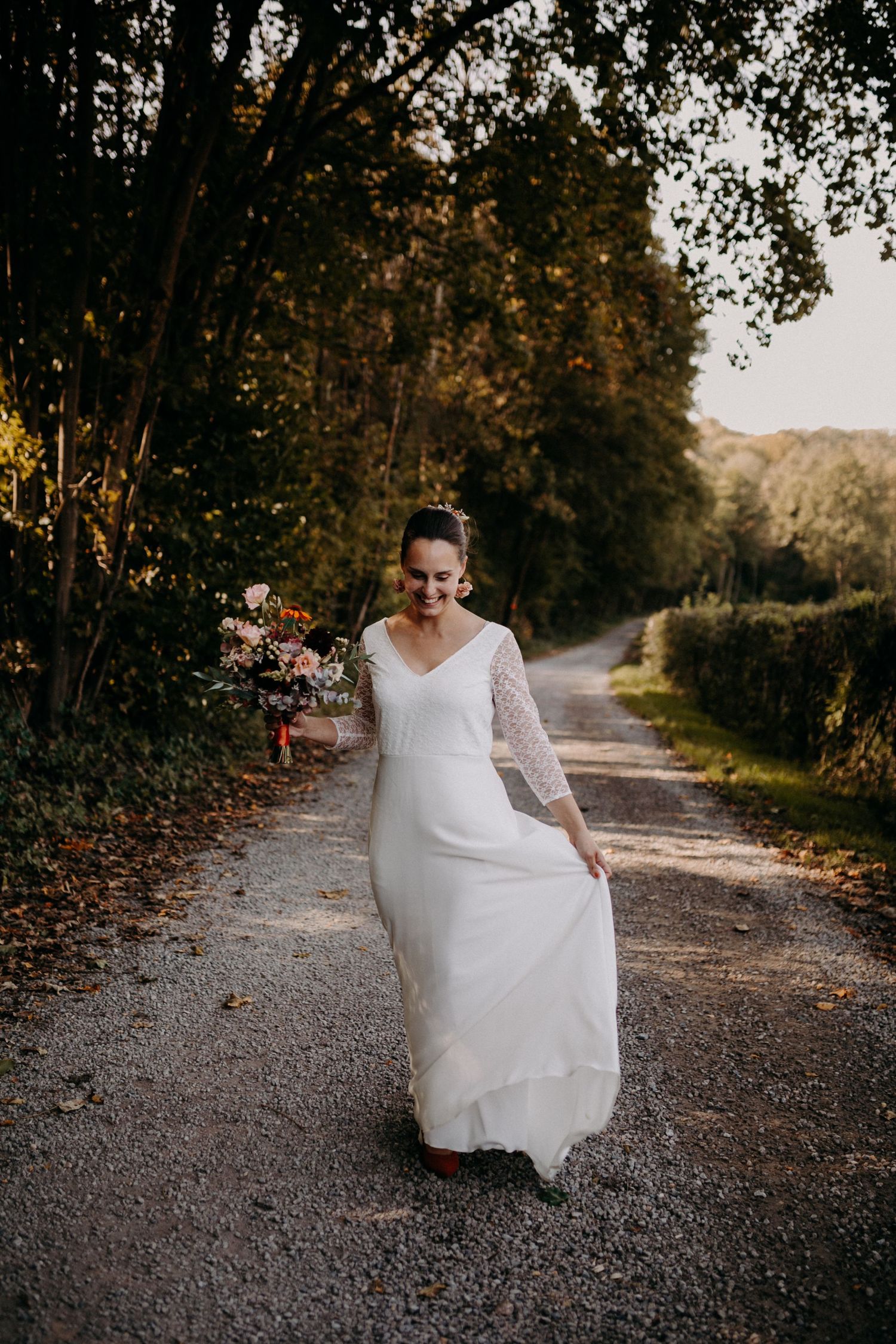 A bride in a long-sleeved white wedding dress walks along a serene autumn path surrounded by trees at sunset.