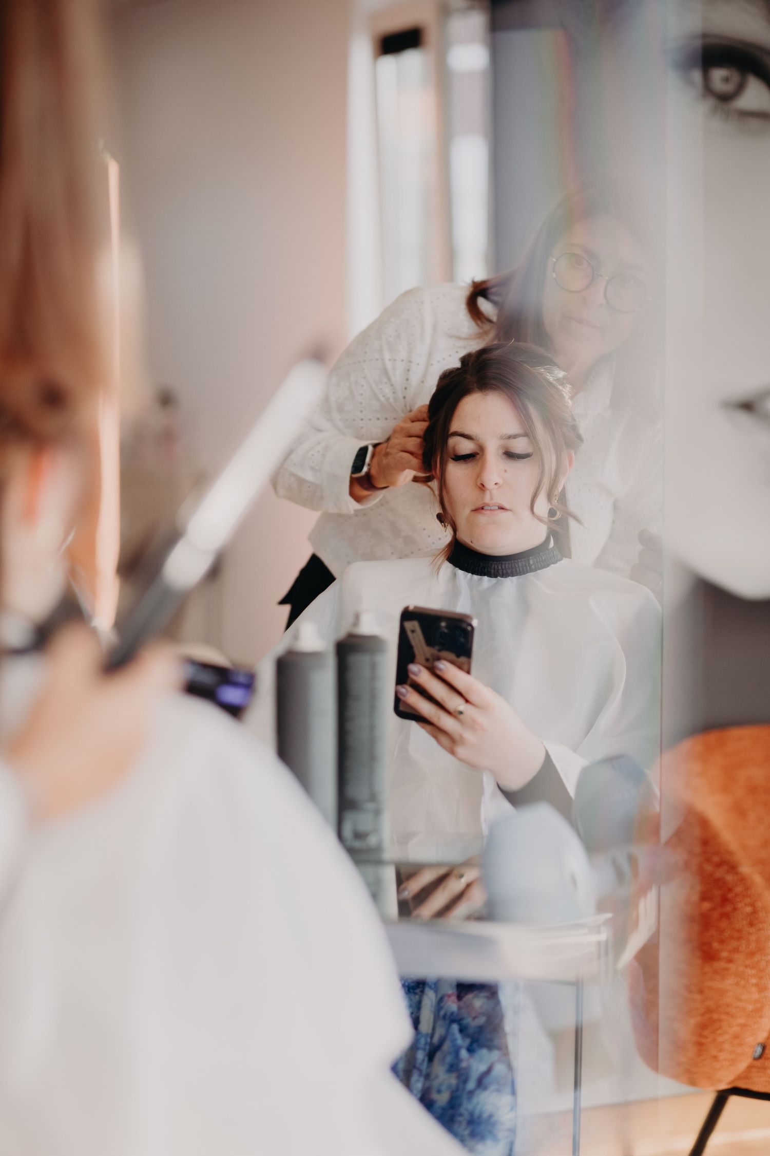 Professional hair stylist styling client's hair while client checks phone in modern salon.
