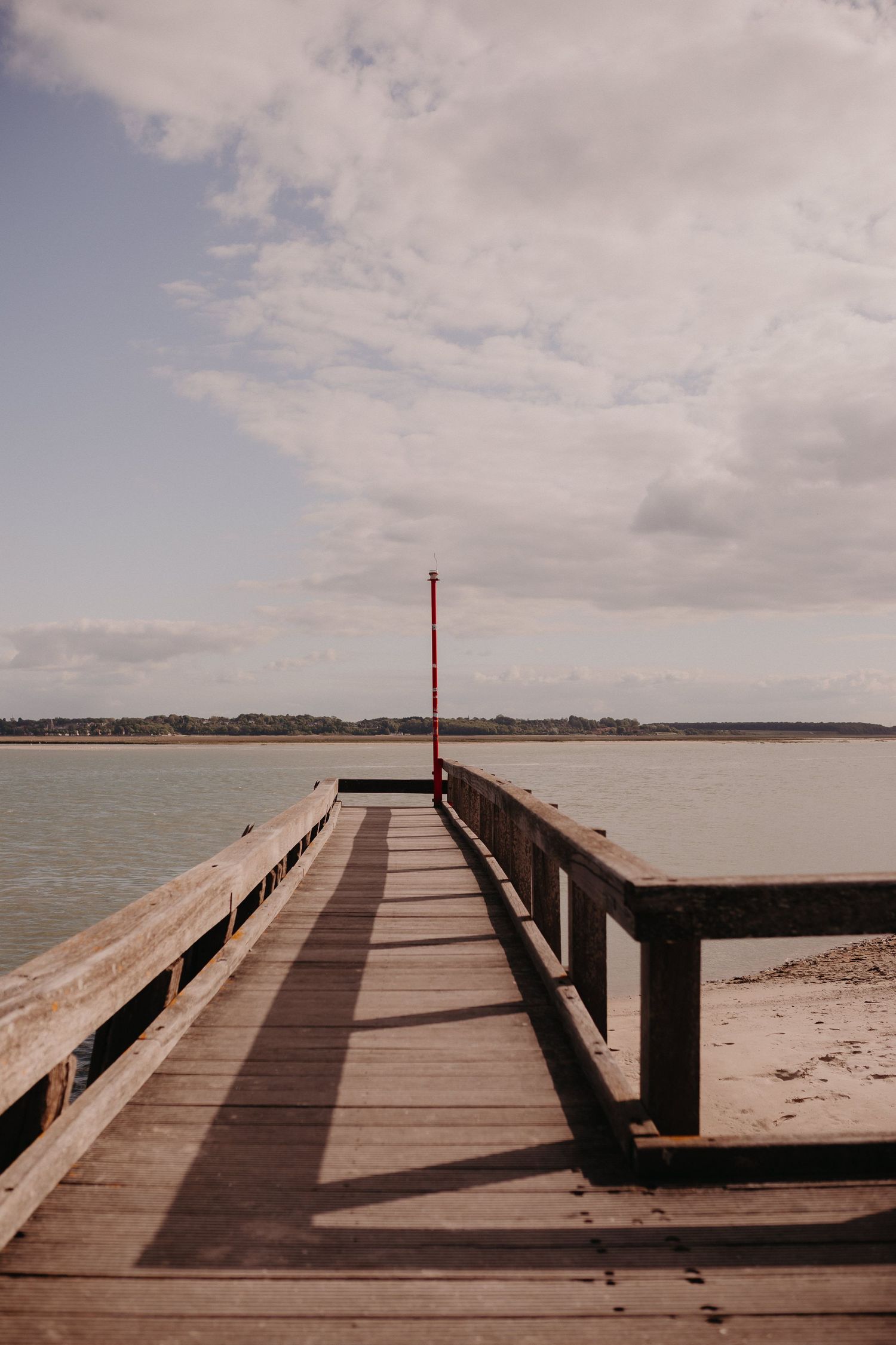 A wooden pier stretches out over calm waters beneath cloudy skies with a single red pole marker at the end.