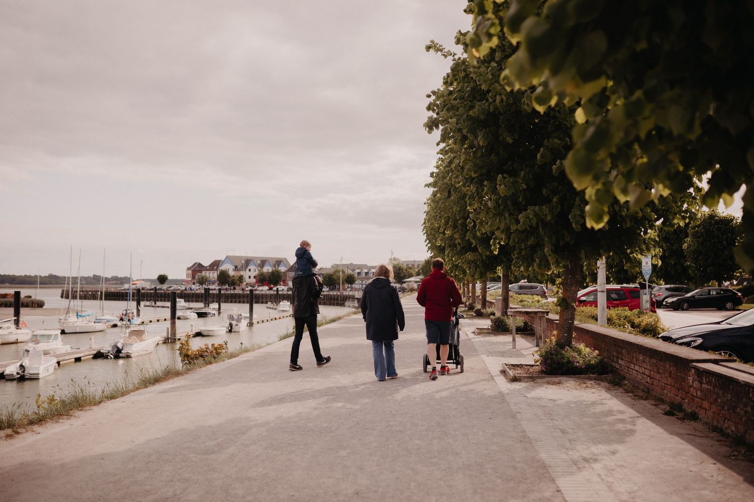 People walking along a waterfront promenade with trees and boats in the marina on an overcast day.