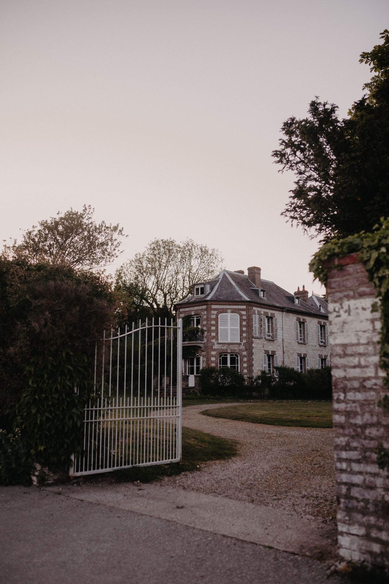 An elegant stone mansion with a curved driveway visible through open metal gates at twilight.
