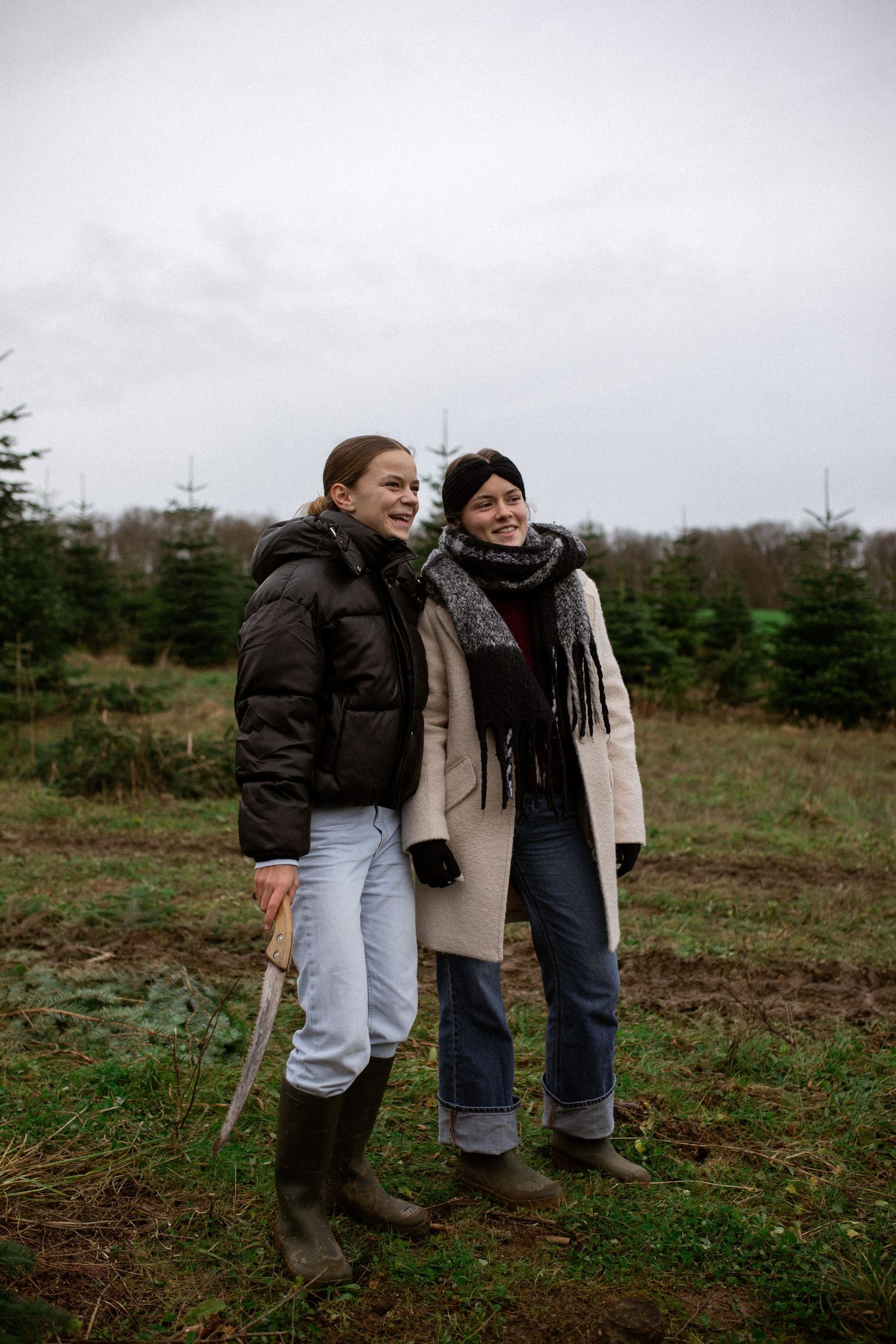 Two people in winter coats and boots stand together in a field with evergreen trees on an overcast day.