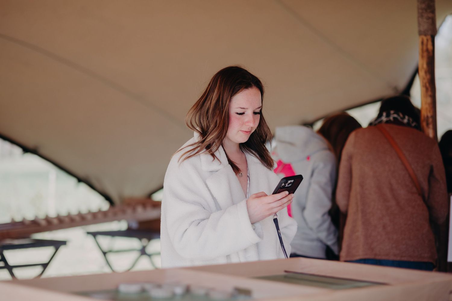 Person in a white coat looking down at mobile phone while standing under a tent or canopy structure.