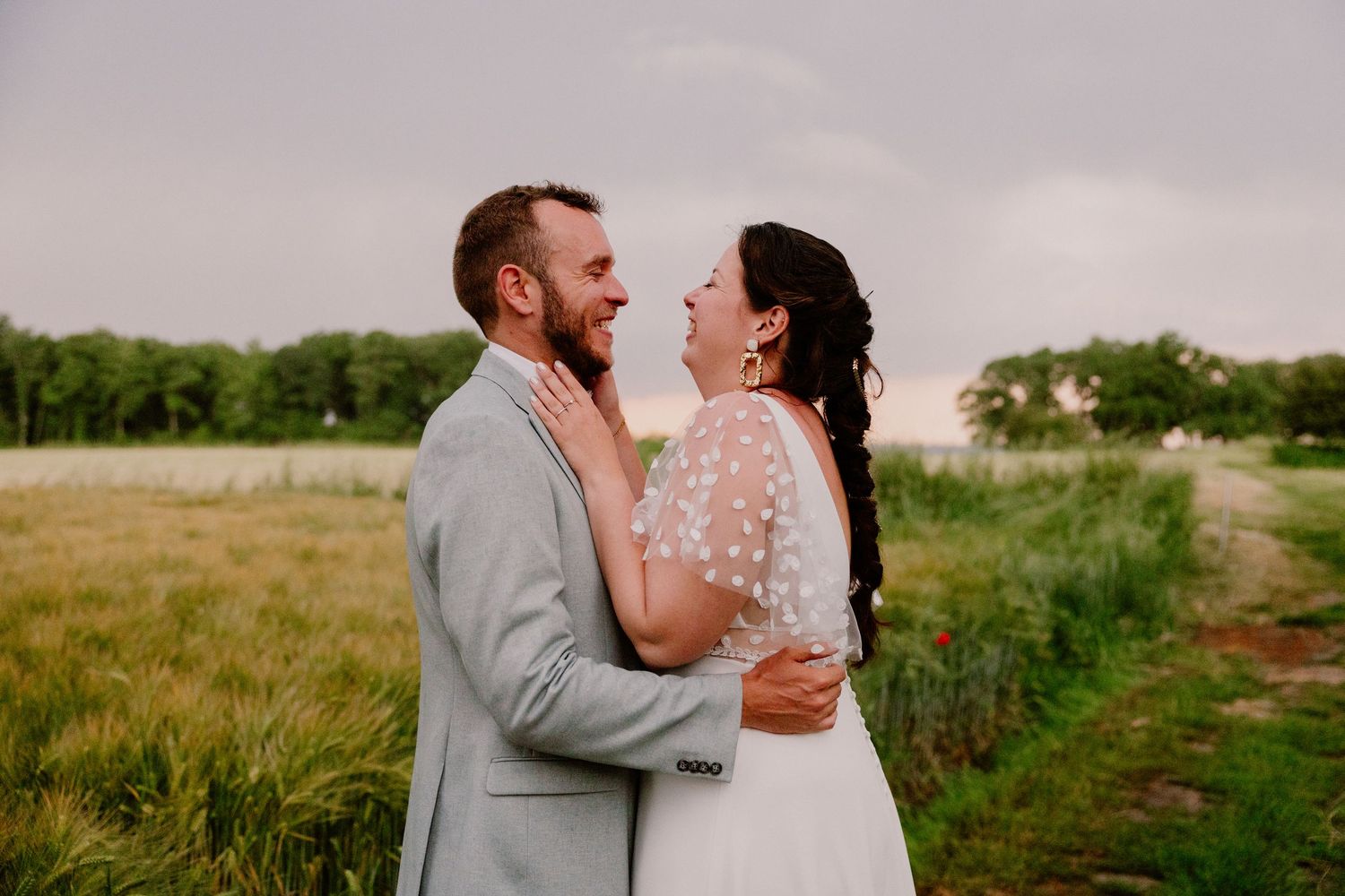 Bride and groom share a joyful moment in a scenic field at sunset, with lush greenery and dramatic skies behind them.