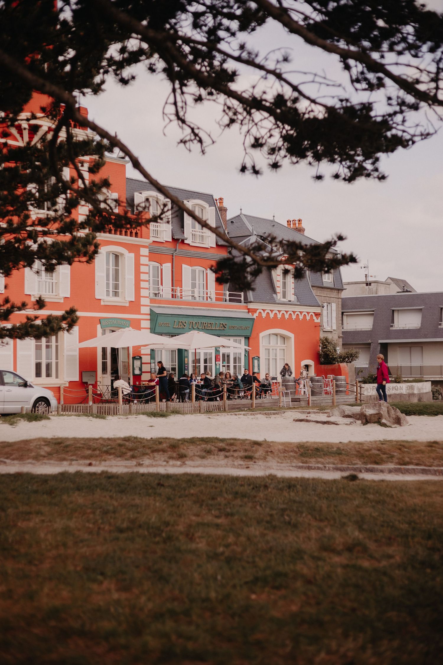 Pink coastal restaurant building with outdoor seating and tree branches framing the view on a cloudy day.