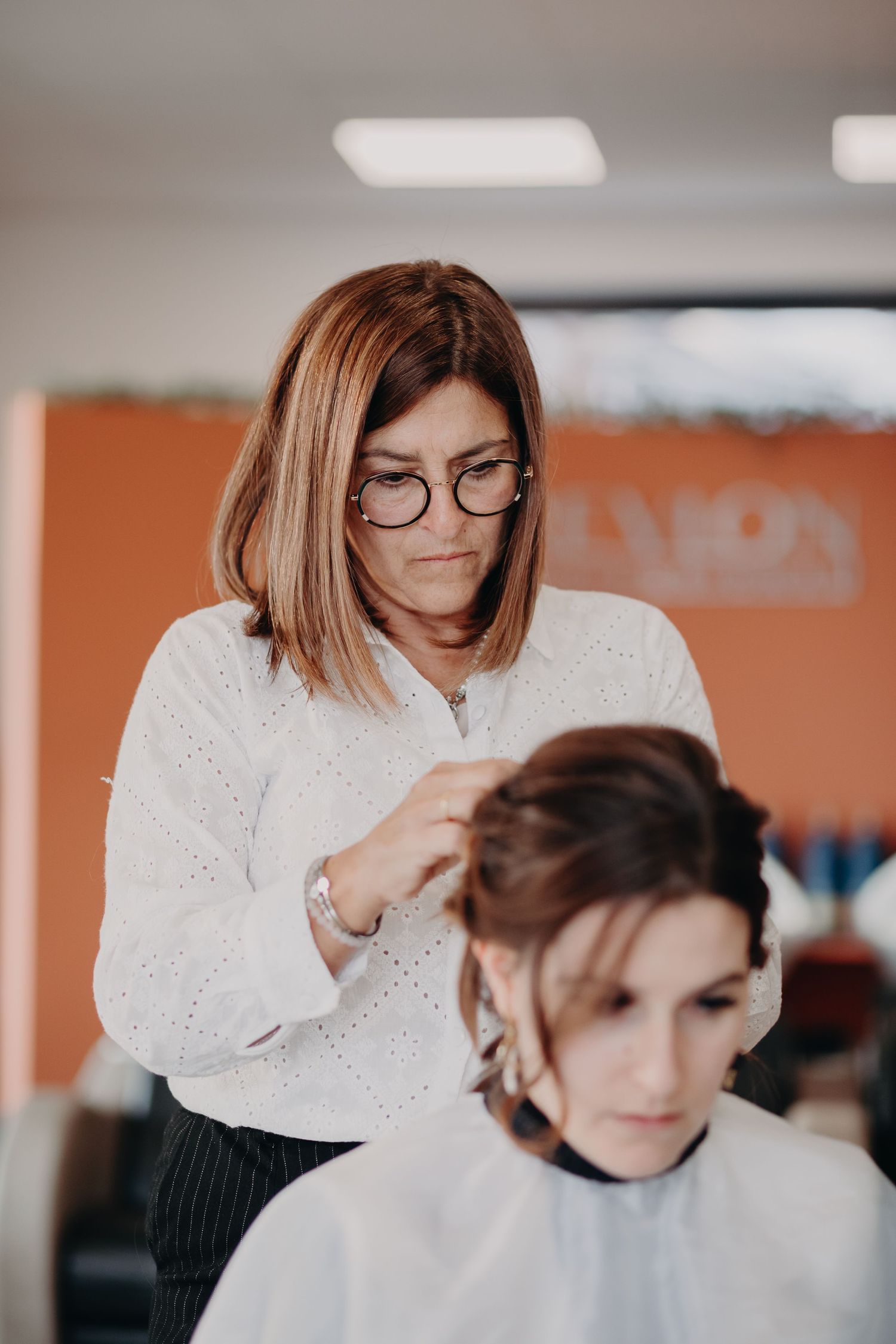 A hairstylist wearing glasses and a white sweater styles a client's hair in a modern salon setting.