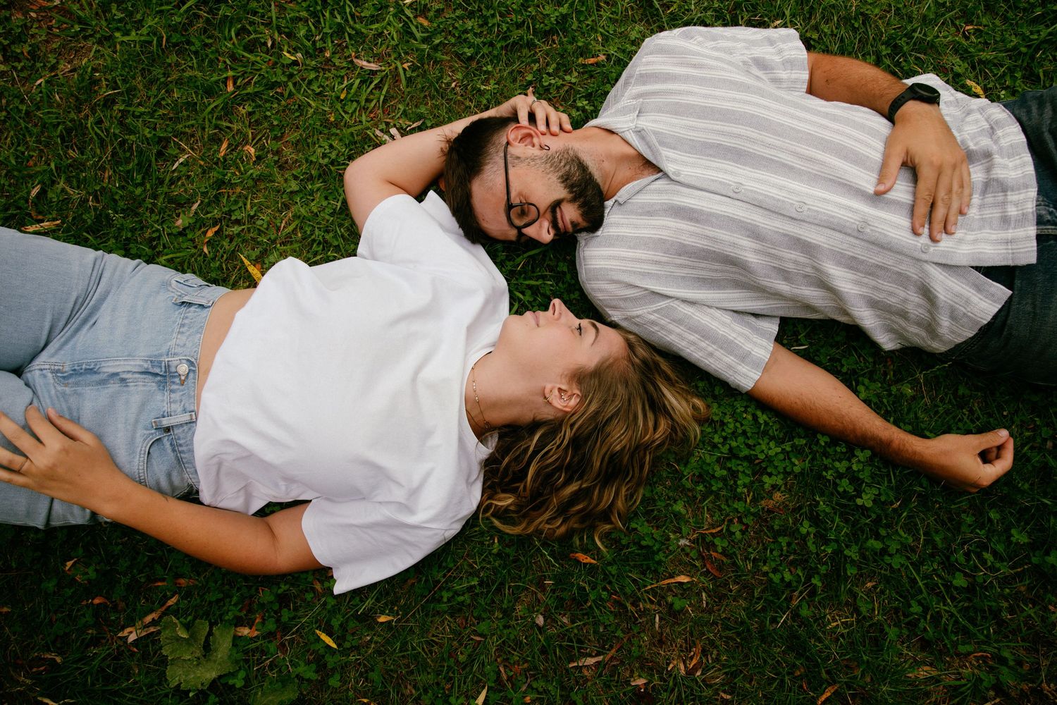 Couple lying on green grass together, sharing a romantic moment outdoors in a park setting.