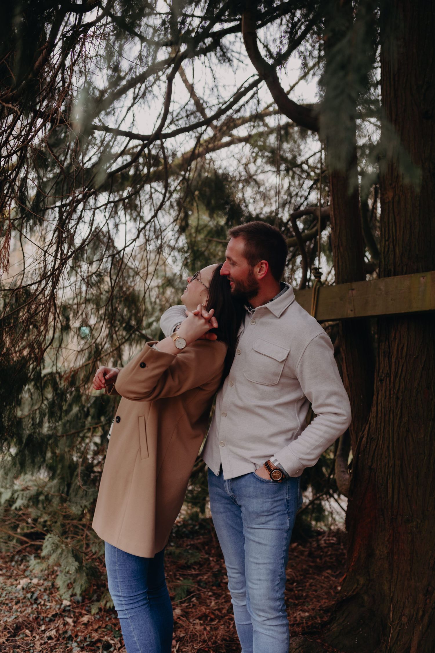 A couple shares a tender moment in nature, leaning against a wooden fence among evergreen trees in moody lighting.
