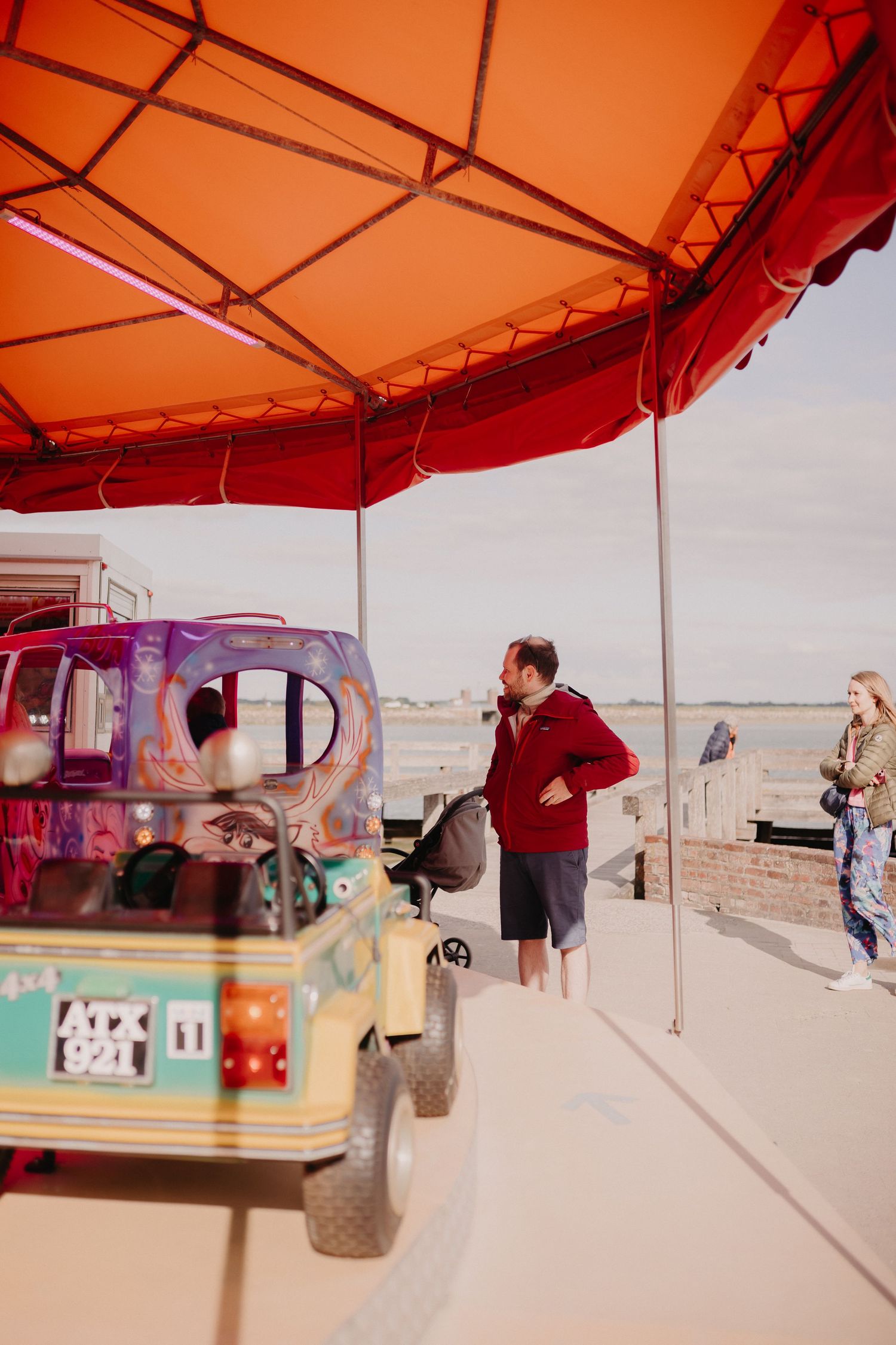 People gather under an orange canopy next to a colorfully decorated vehicle on a desert terrain.