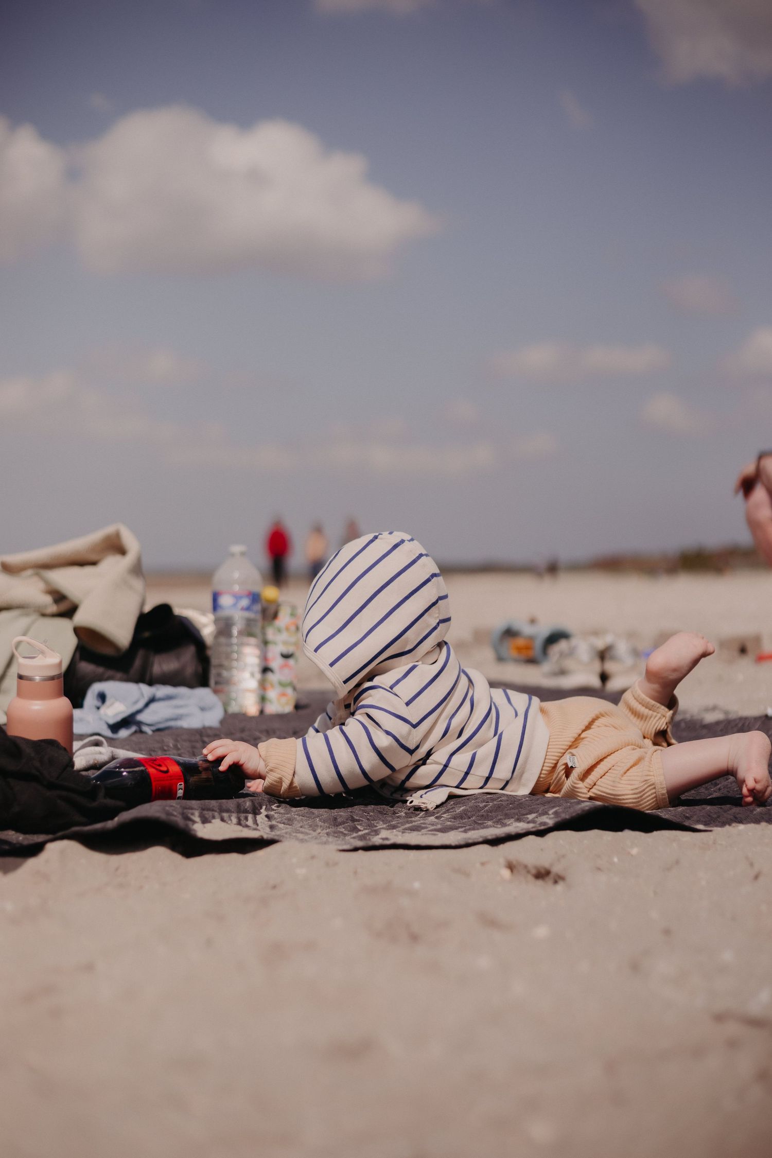 Small child in striped outfit lying on beach blanket relaxing on sandy shore during peaceful summer day.