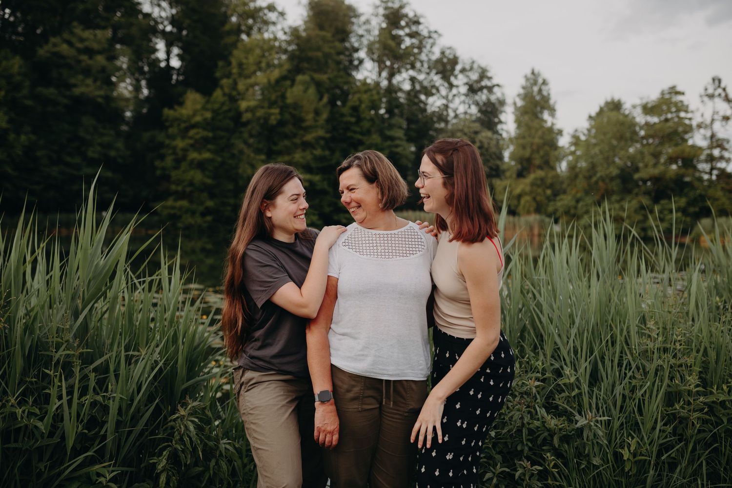 Three women wearing casual outfits stand together and smile while surrounded by tall green grasses in a natural setting.