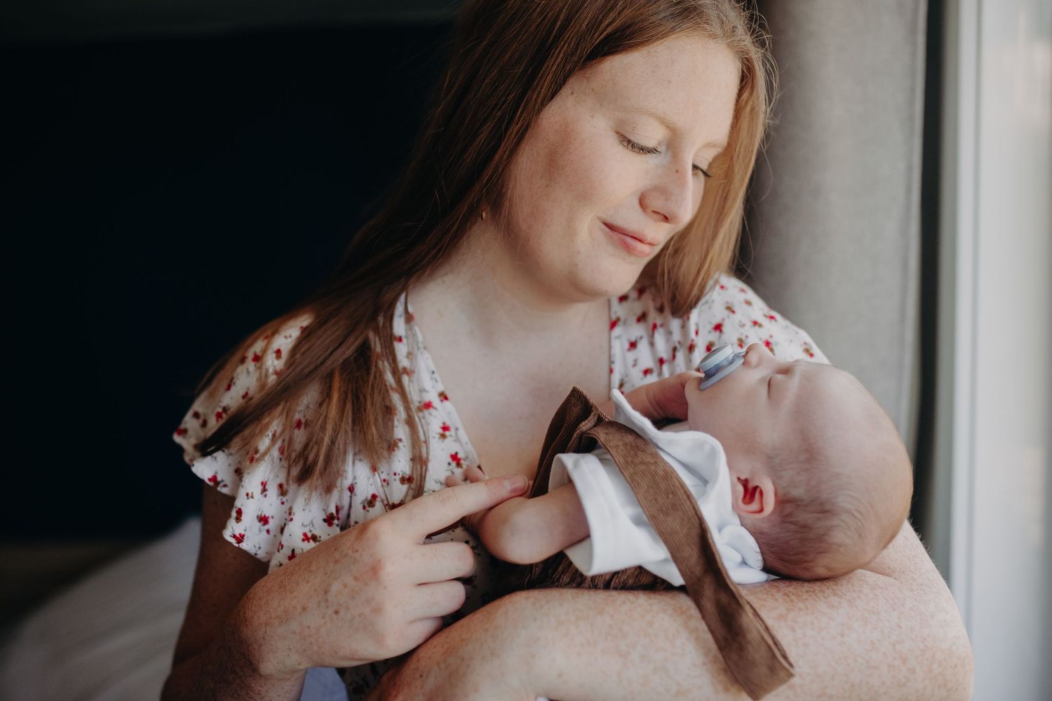 A mother in a floral dress tenderly holds and gazes at her newborn baby in a natural light setting.