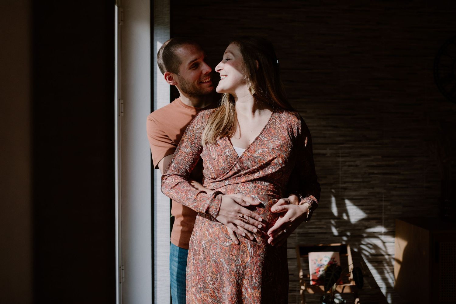 A couple embraces in romantic shadows against a brick wall, wearing casual attire and floral print dress.