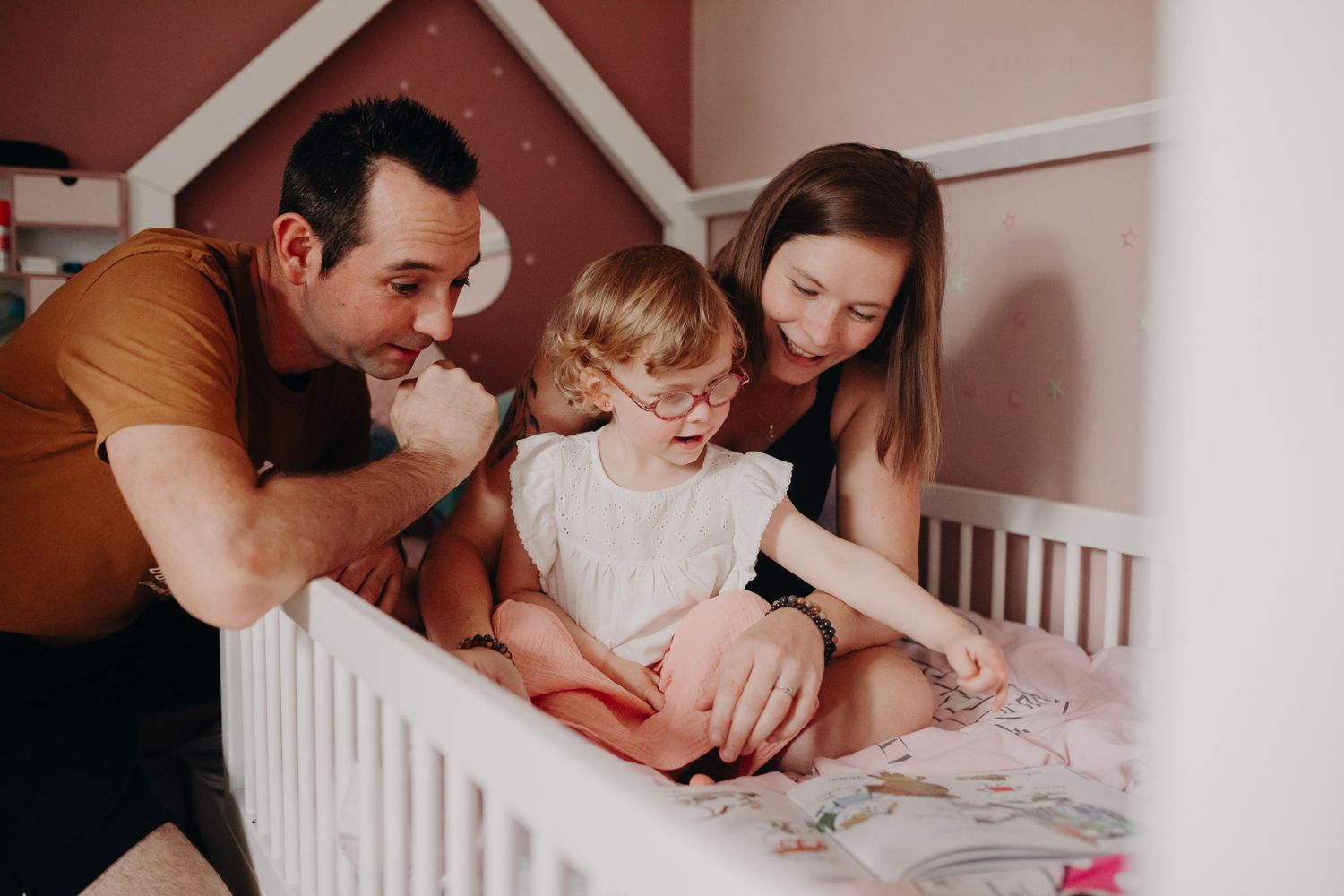 Parents sit with child at white crib while reading a book together.