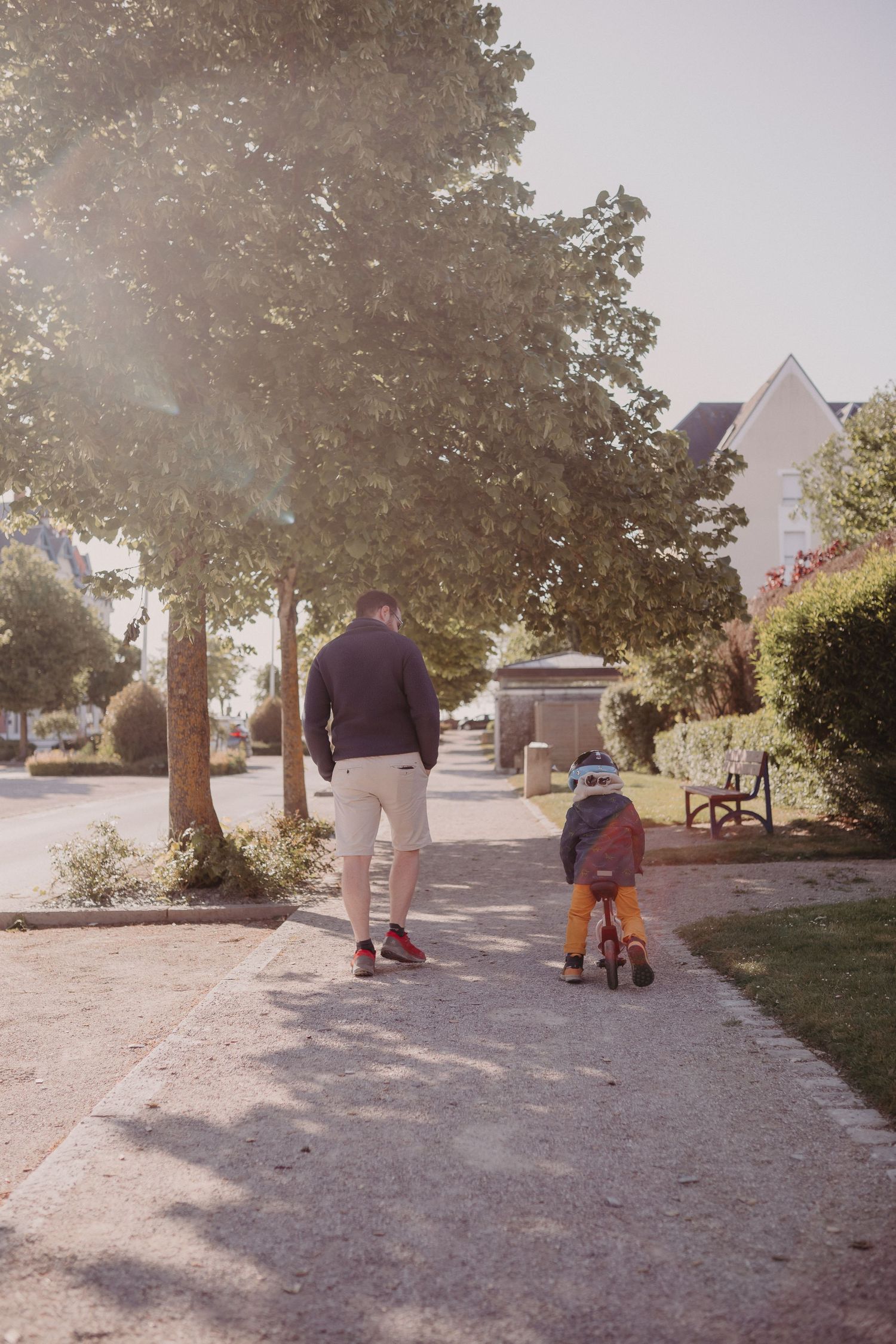 A parent and child walk together on a residential path during a sunny afternoon, with the child riding an orange bicycle.