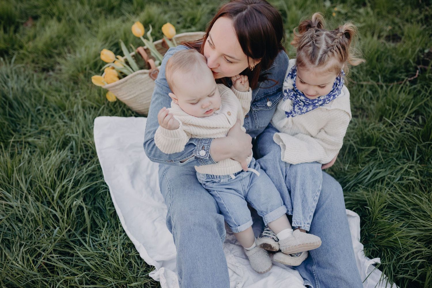 A parent sits with two young children on a white blanket in grassy field during an outdoor family photo session.