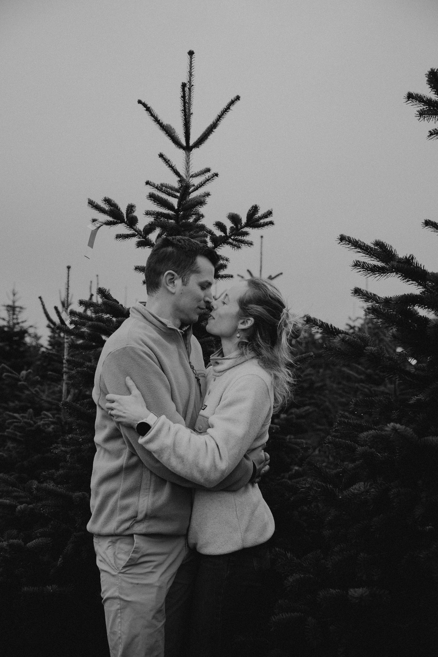 A romantic black and white photo series showing a couple embracing in front of evergreen trees.