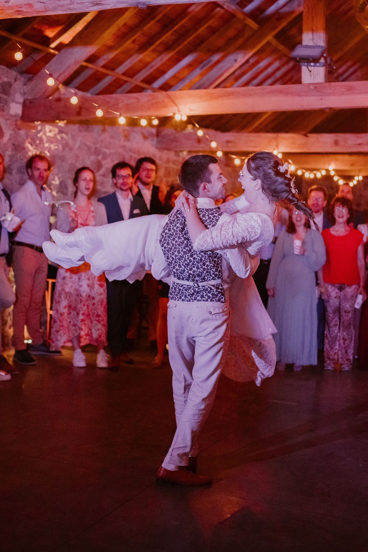 A couple dances at a rustic barn reception with string lights overhead and guests watching in the background.