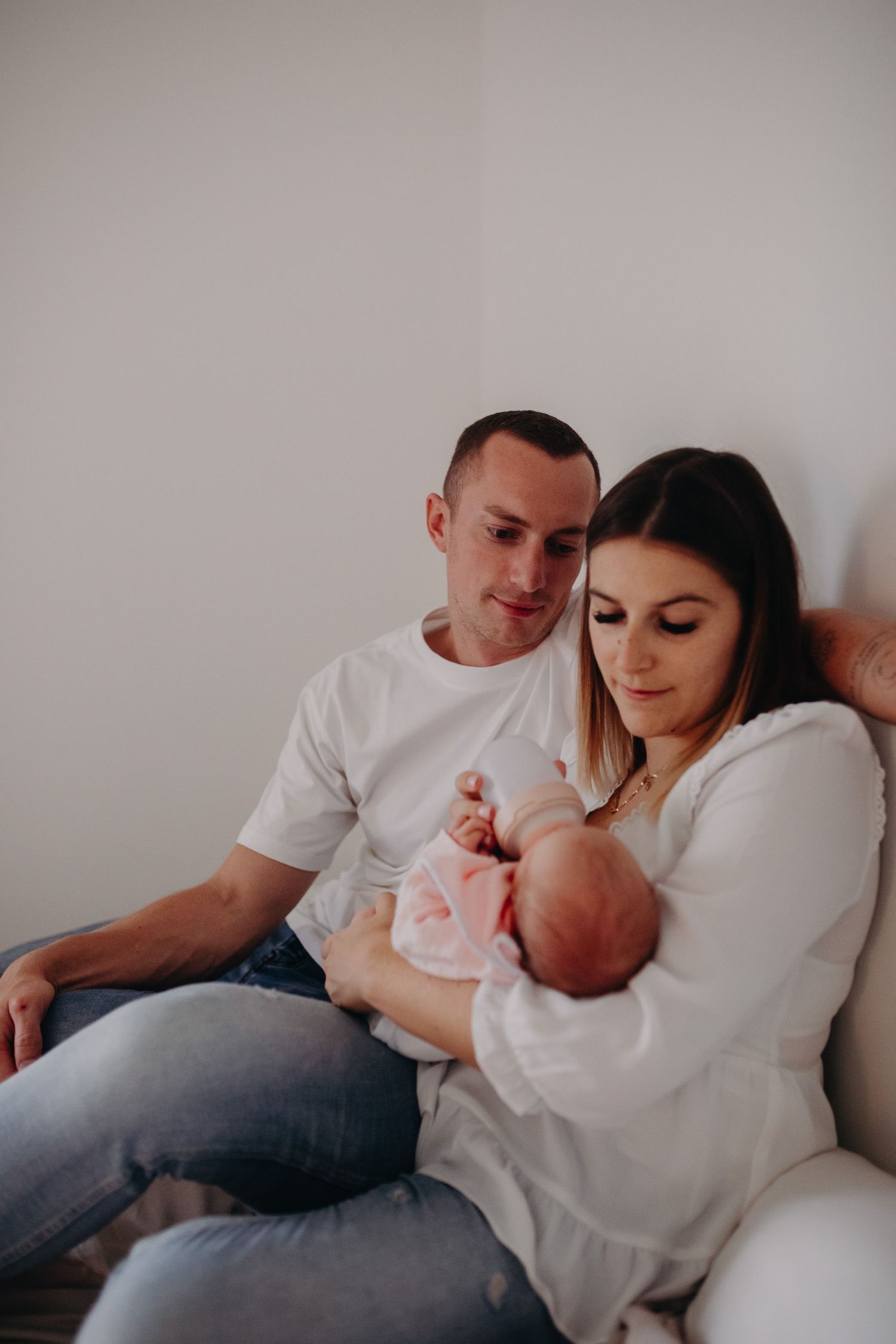 A couple sits together on a couch while tenderly gazing at their newborn baby in a warm, intimate home setting.