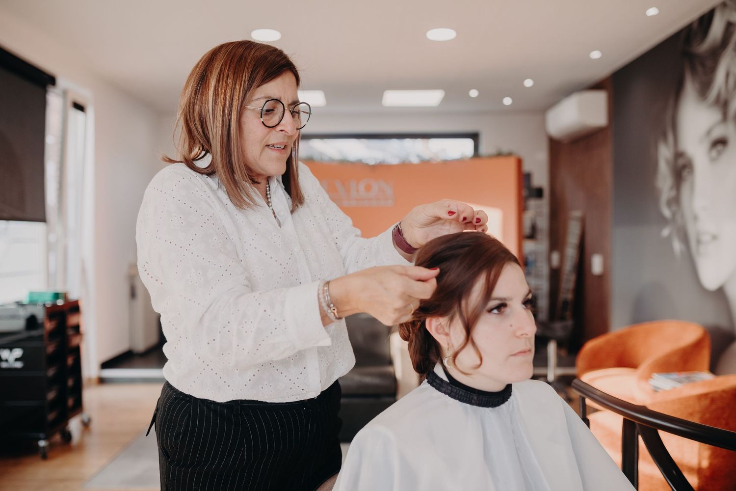 A hairstylist wearing glasses and a white sweater styles a client's hair in a modern salon with orange accents.