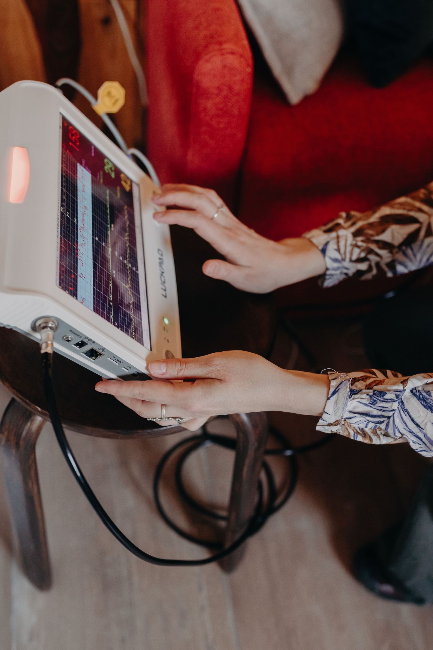 Hands holding an electronic tablet device with cables attached, wearing patterned sleeves against a red background.
