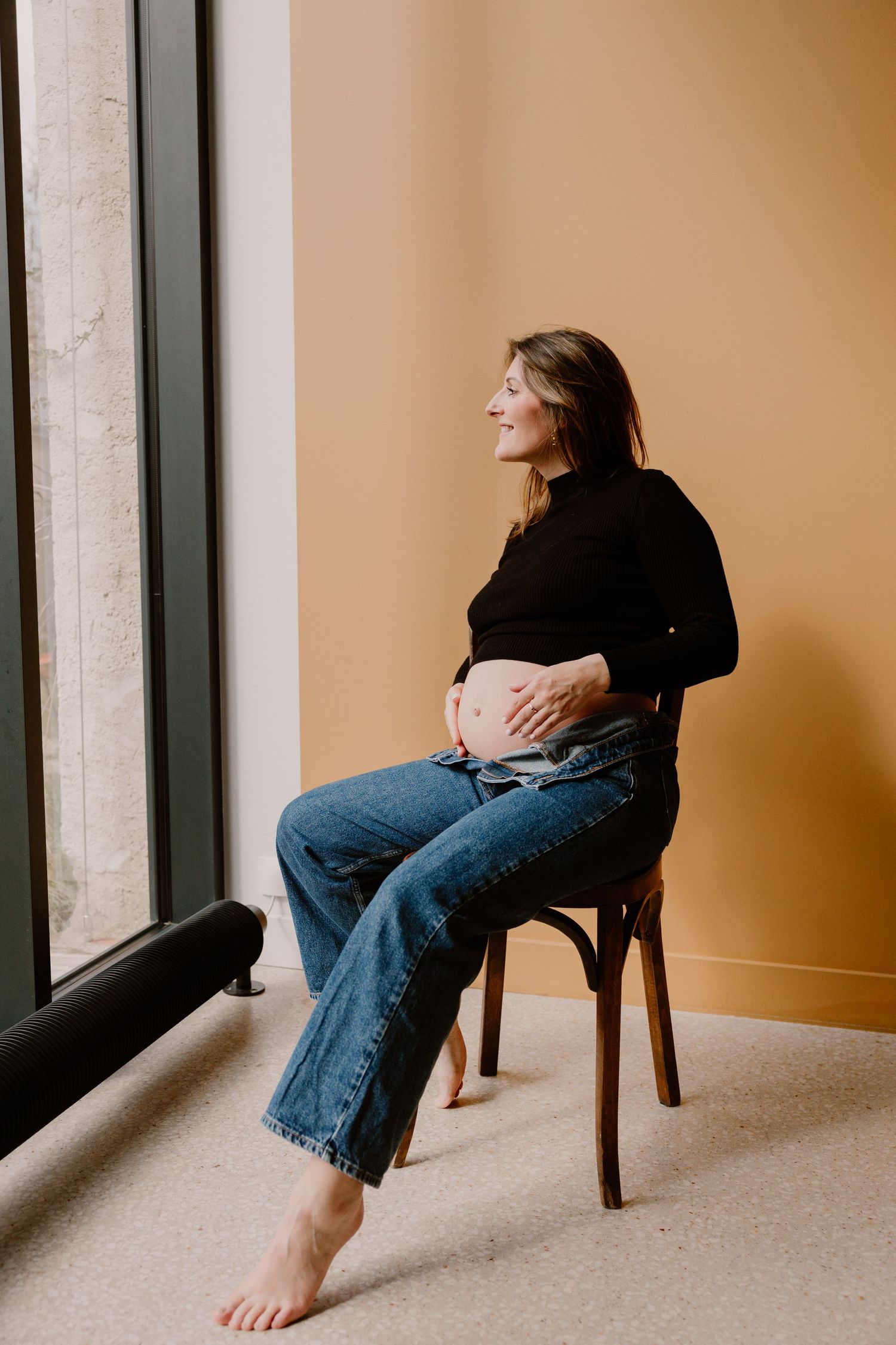 Smiling pregnant woman sitting on wooden chair touching baby bump, wearing black turtleneck and jeans near window.