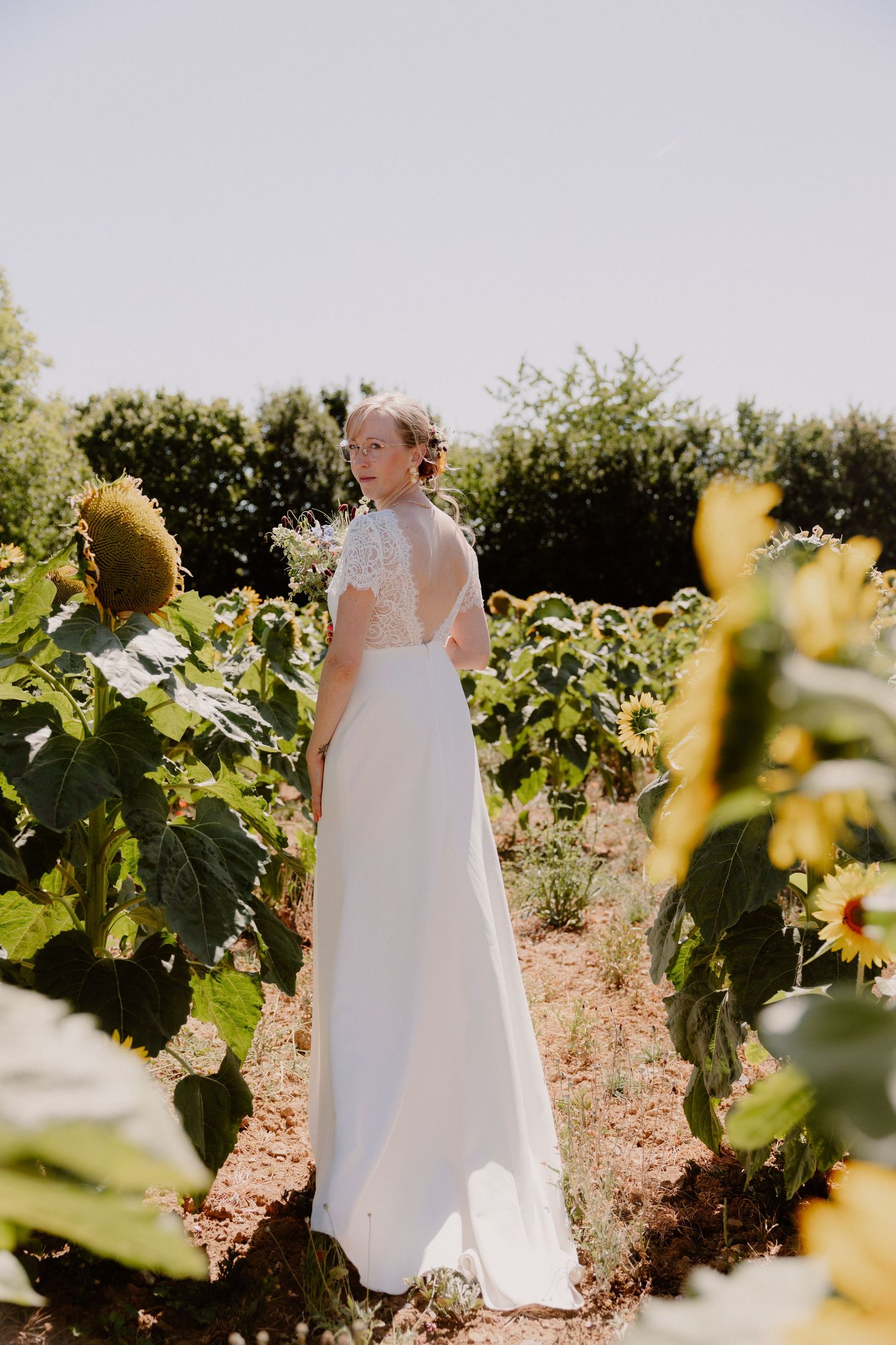 Bride in white lace gown standing among sunflowers in a sunny garden, viewed from behind.