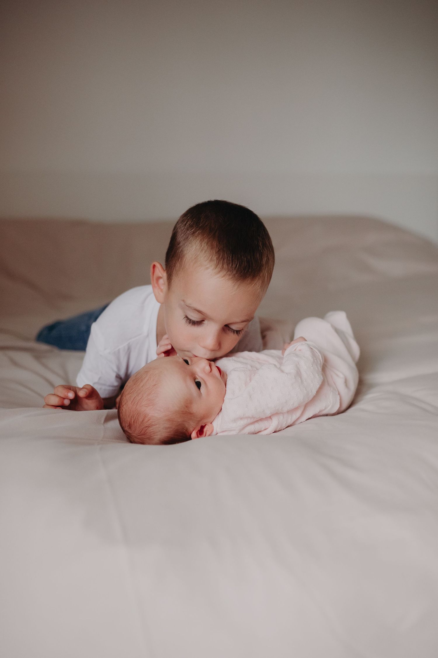 Two children share a tender moment lying together on white bedding in a softly lit room.