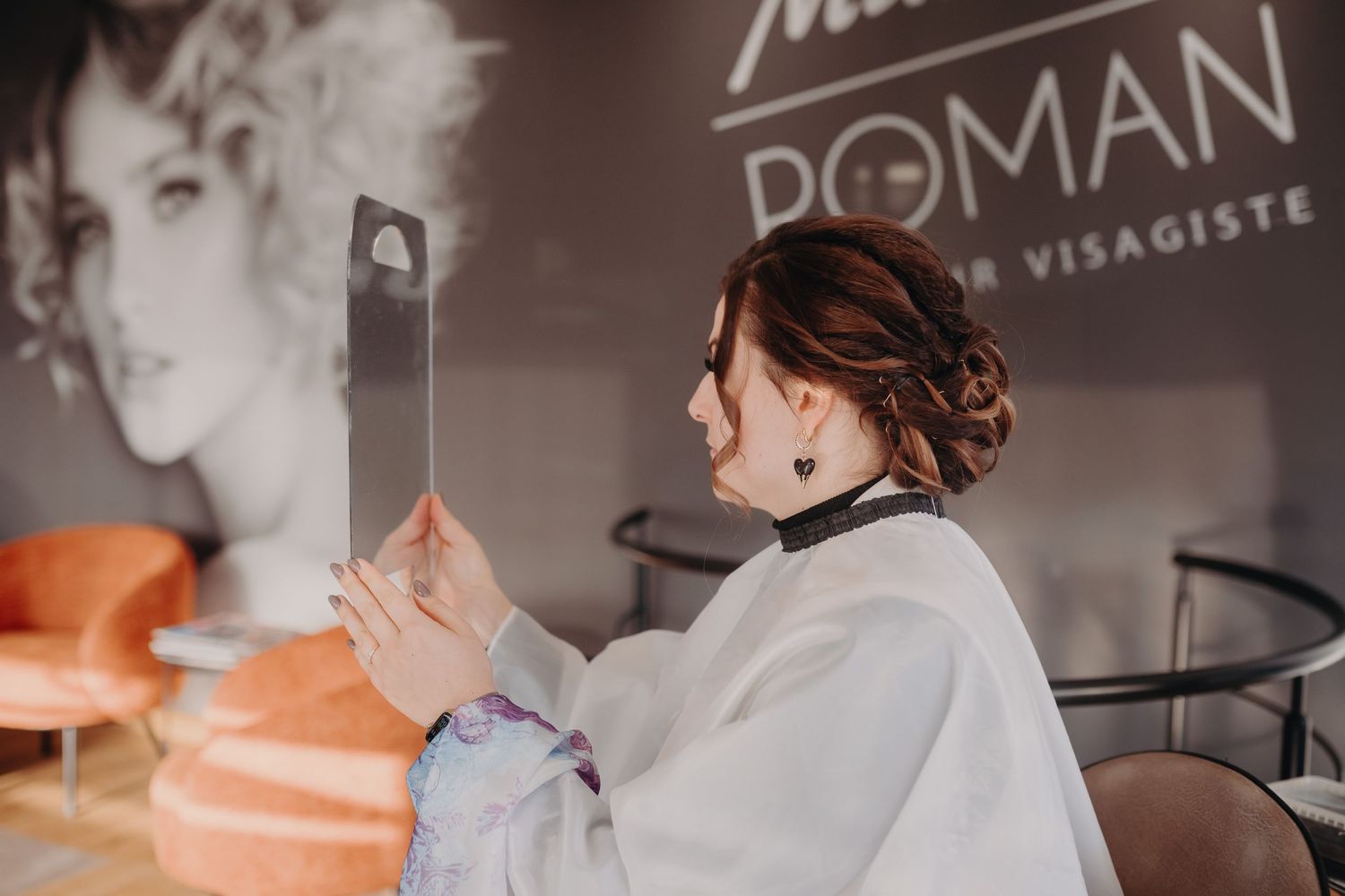 Hairstylist wearing white coat takes phone photo of work in salon with retro decor and branding visible.