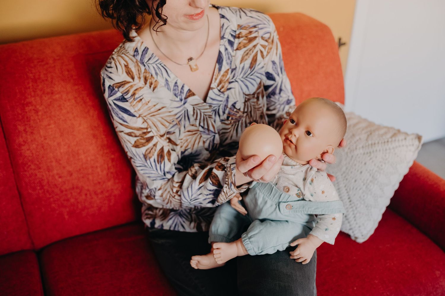 A person in a floral print top sits on a red couch while holding and feeding an infant in light blue clothing.