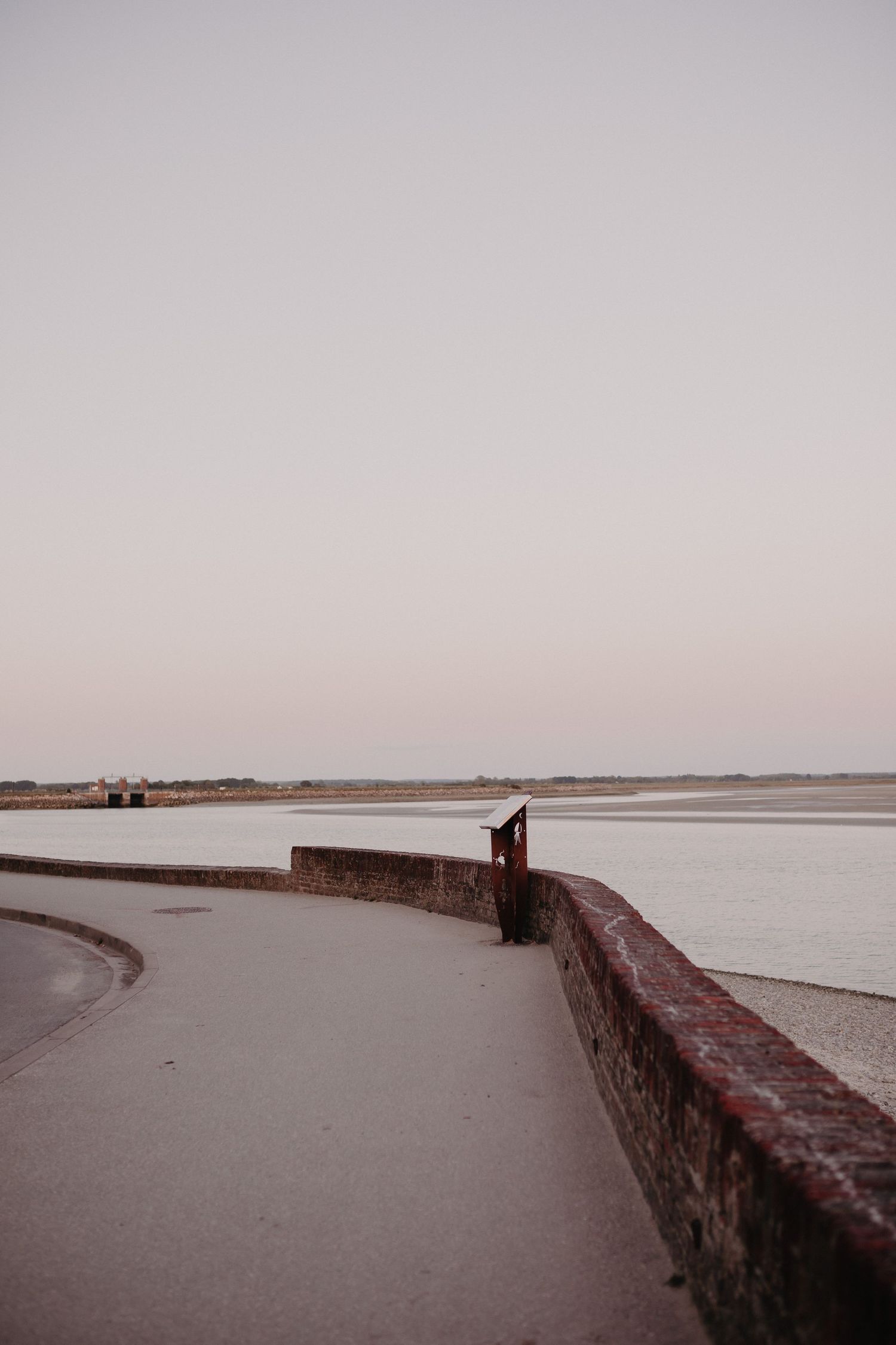 A curved concrete walkway with red brick wall follows along a calm coastline at dusk.