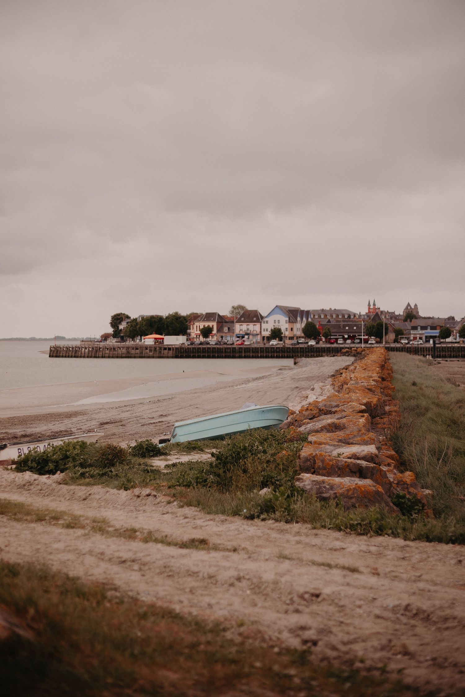Moody coastal landscape with overturned boat and seaside houses along a rocky breakwater under cloudy skies.