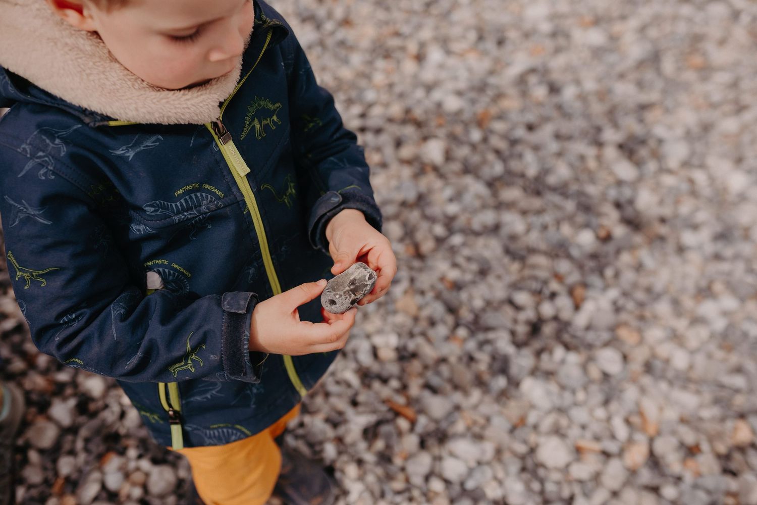 A person wearing a navy blue jacket and yellow socks examines something in their hands while standing on pebbles.