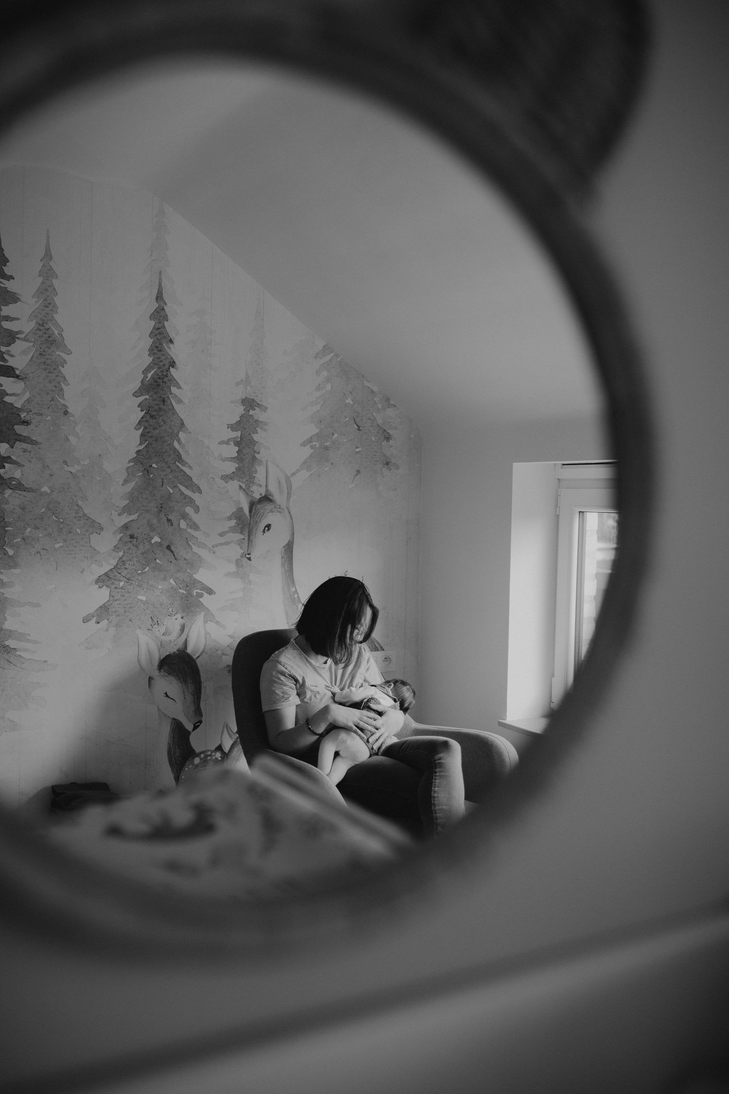 Black and white photo viewed through a circular frame showing a nursery with pine tree wall mural and figure in rocking chair.