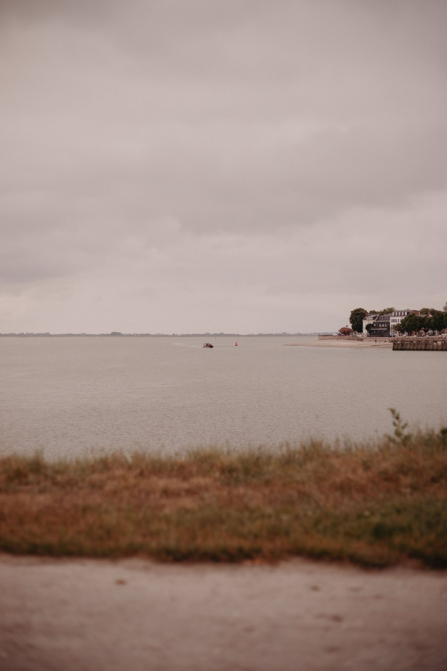 A cloudy coastal landscape with brown grass in foreground and buildings visible along the waterfront on a moody day.