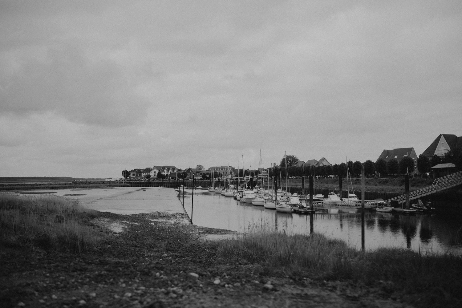 A black and white waterfront marina with moored boats and houses along the shoreline under cloudy skies.
