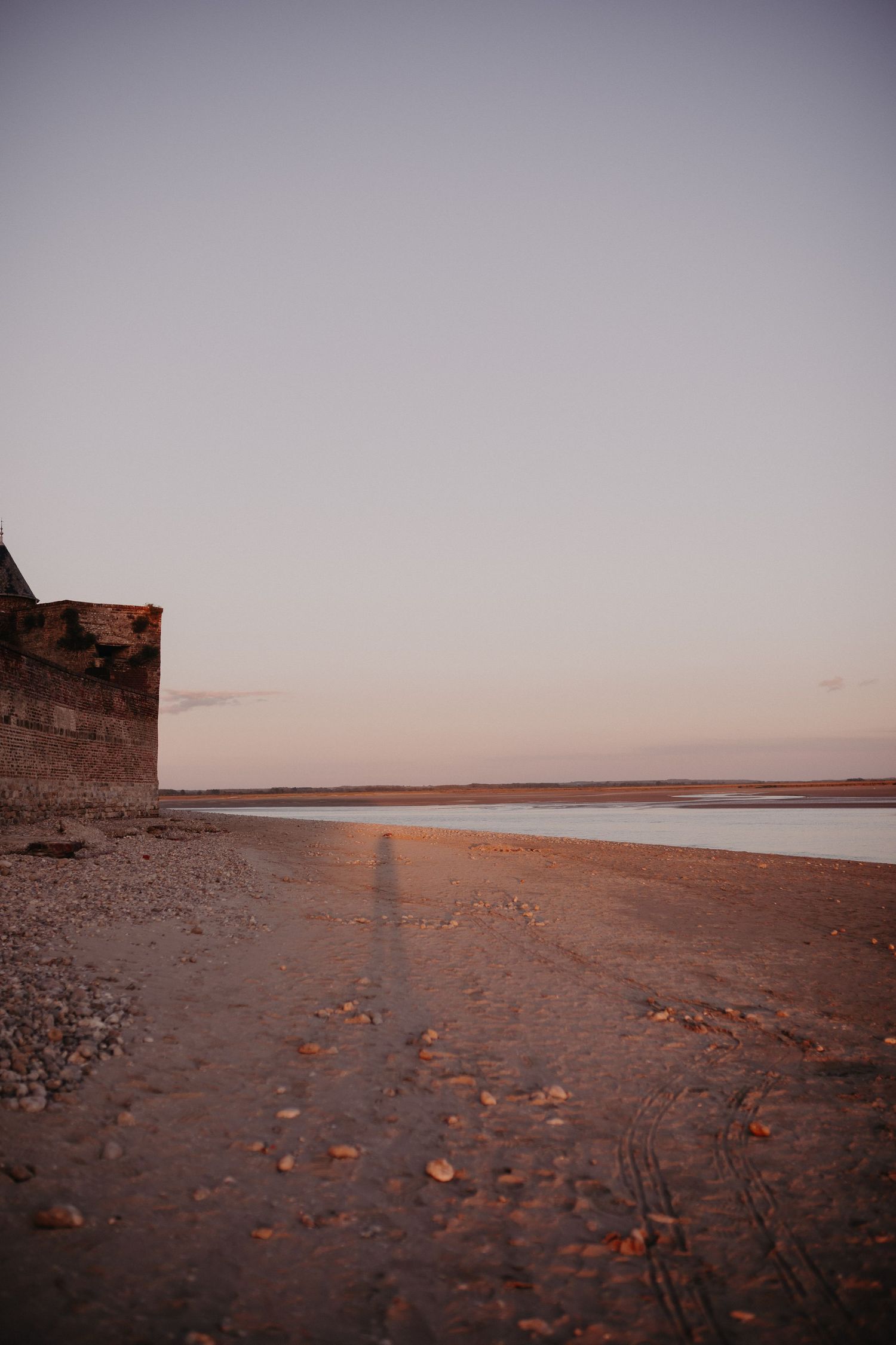 A sandy beach at sunset with a stone wall and long shadow cast across the shore during golden hour.