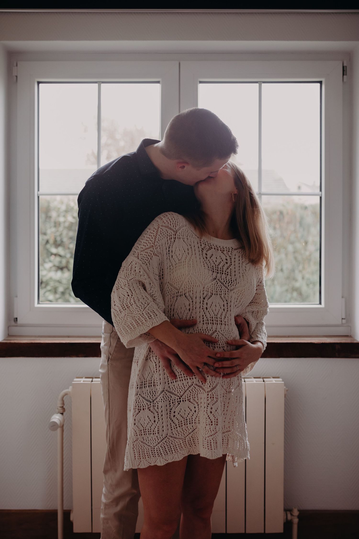 A romantic moment shared between a couple by a window, with the person wearing a cream knit dress and the other in dark attire.