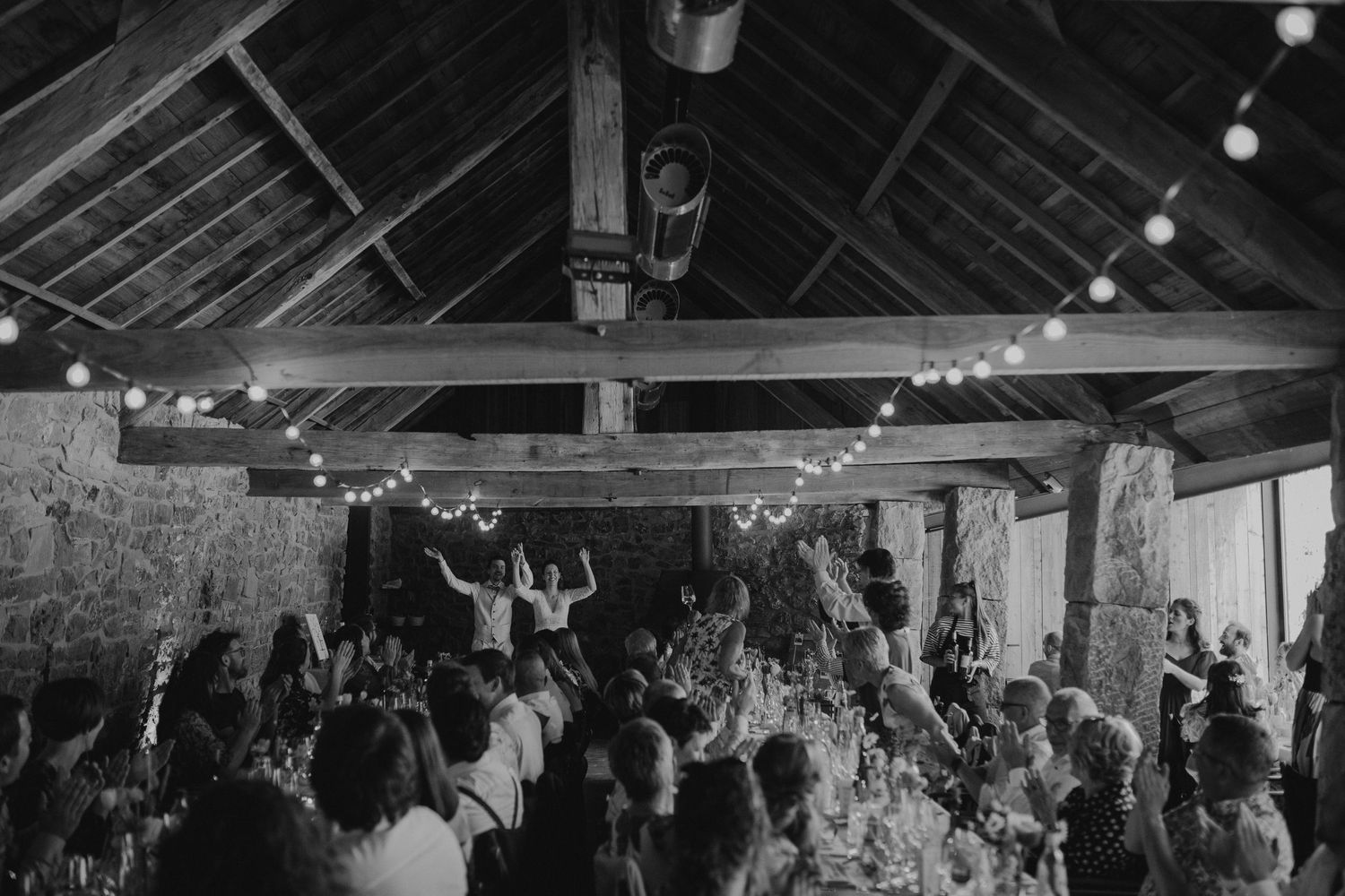 A crowded party or celebration in a rustic barn with string lights hung from wooden beams overhead.