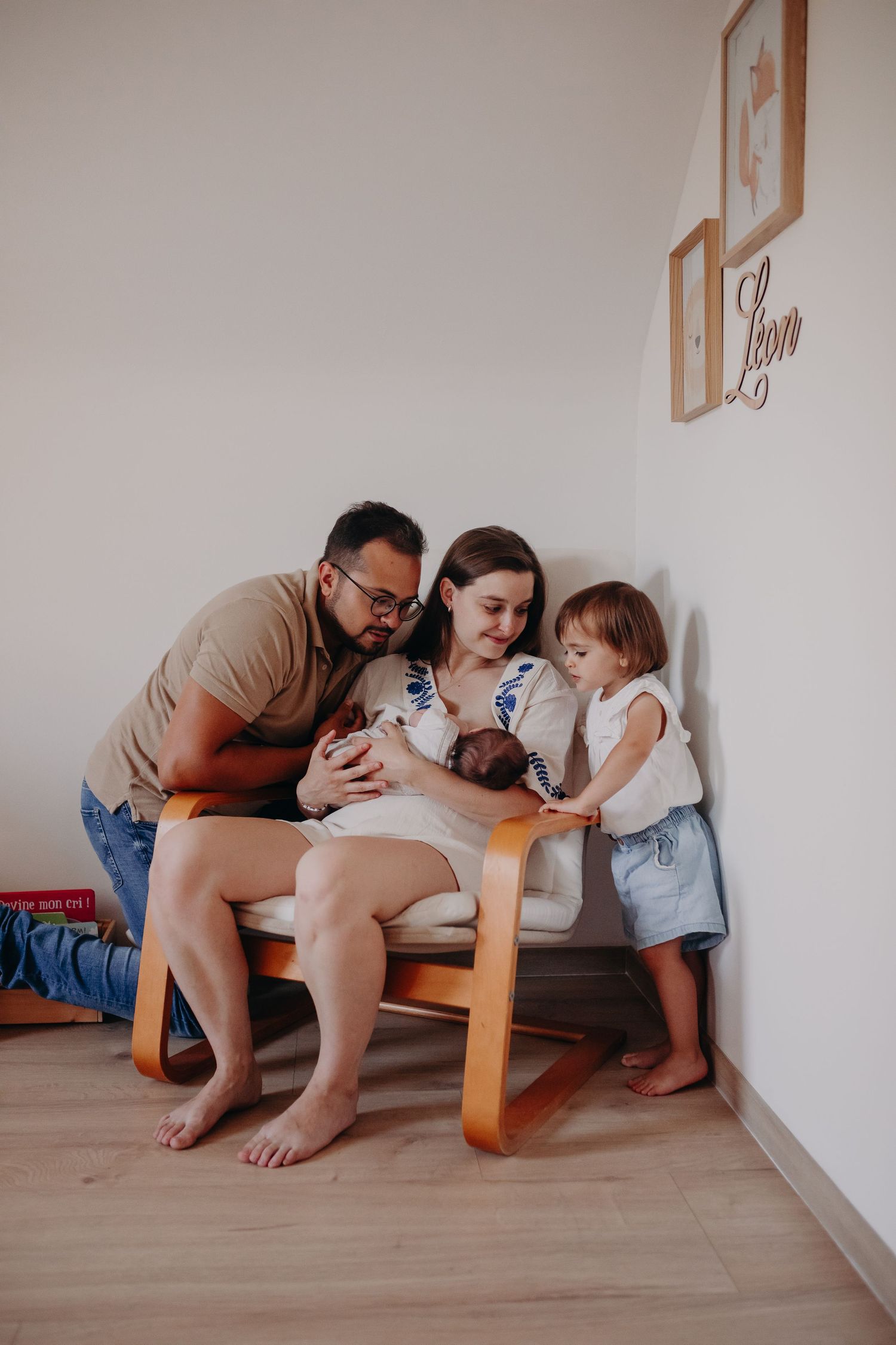 Family relaxes together on a wooden chair in a minimalist room with light walls and hardwood flooring.