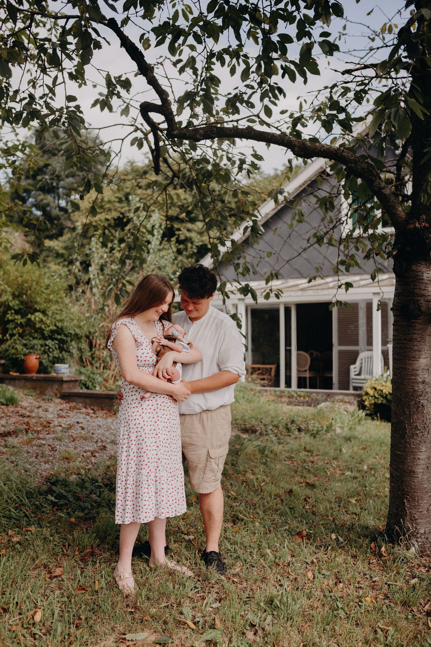 A couple shares a tender moment under a tree in a backyard setting with a white house in the background.
