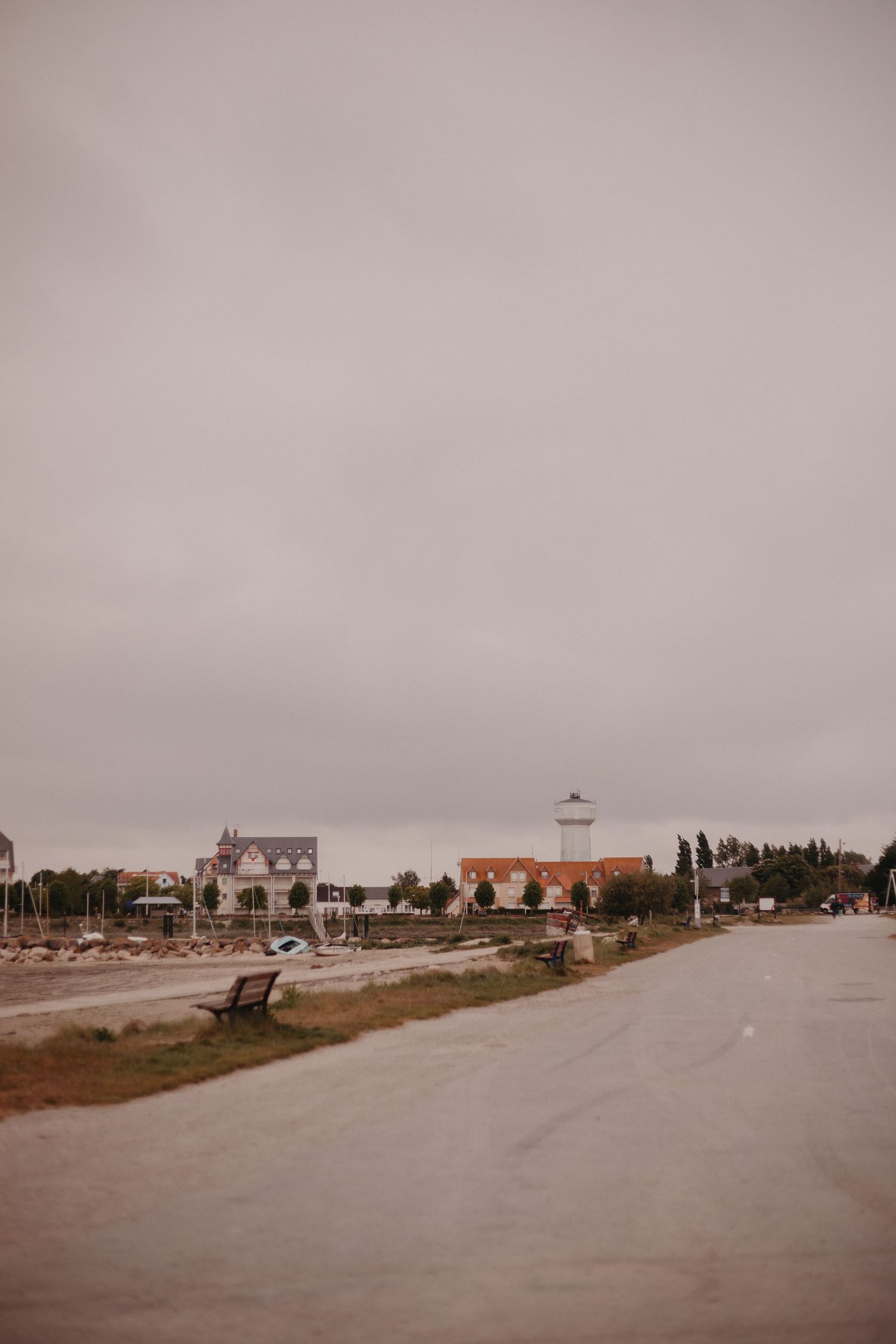 Coastal road leading to a quaint seaside village with white buildings and lighthouse under an overcast sky.