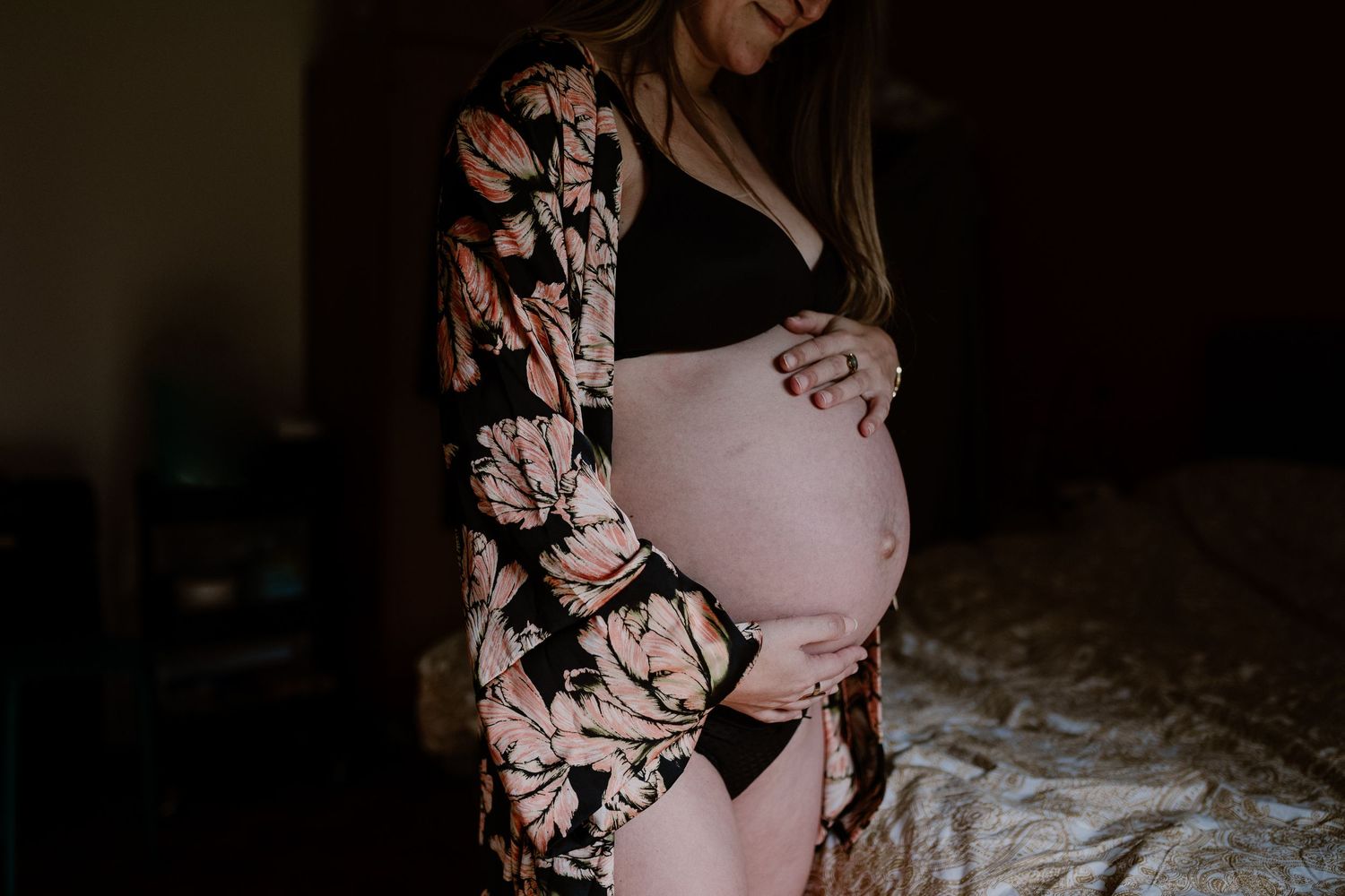 A person in a floral robe touches their pregnant belly in moody lighting.