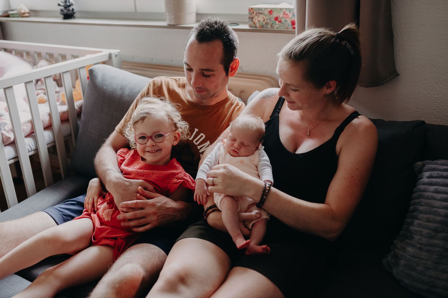 Family sitting together on cozy couch at home with toddler in red shirt and infant baby.