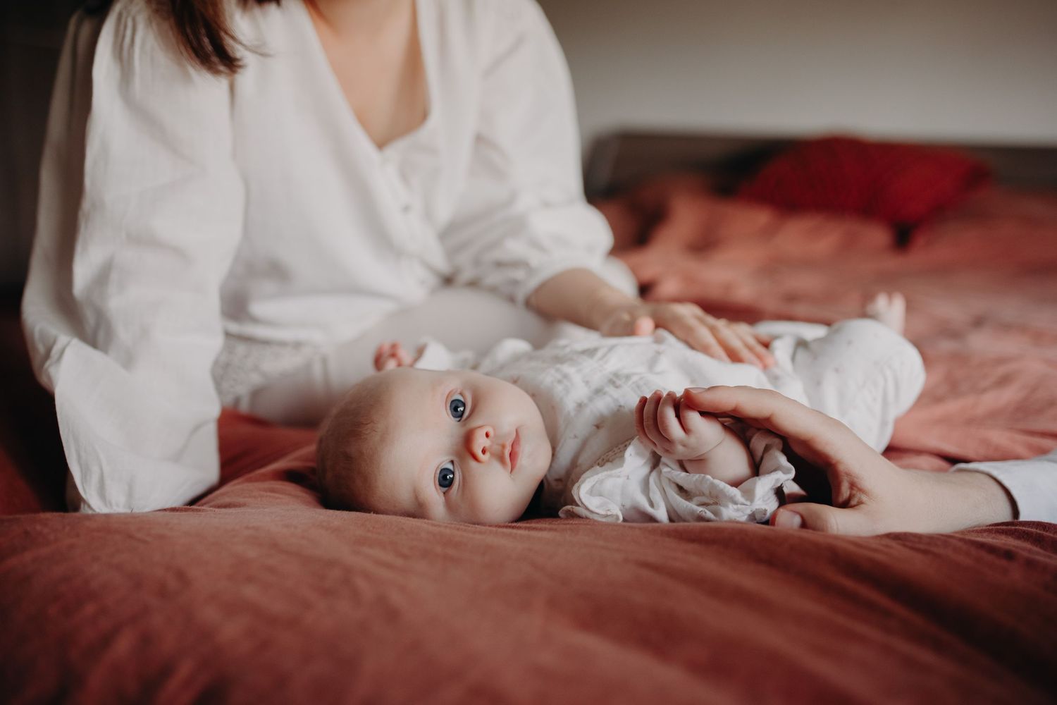A baby dressed in white sleeping peacefully on burgundy bedding while someone sits nearby.