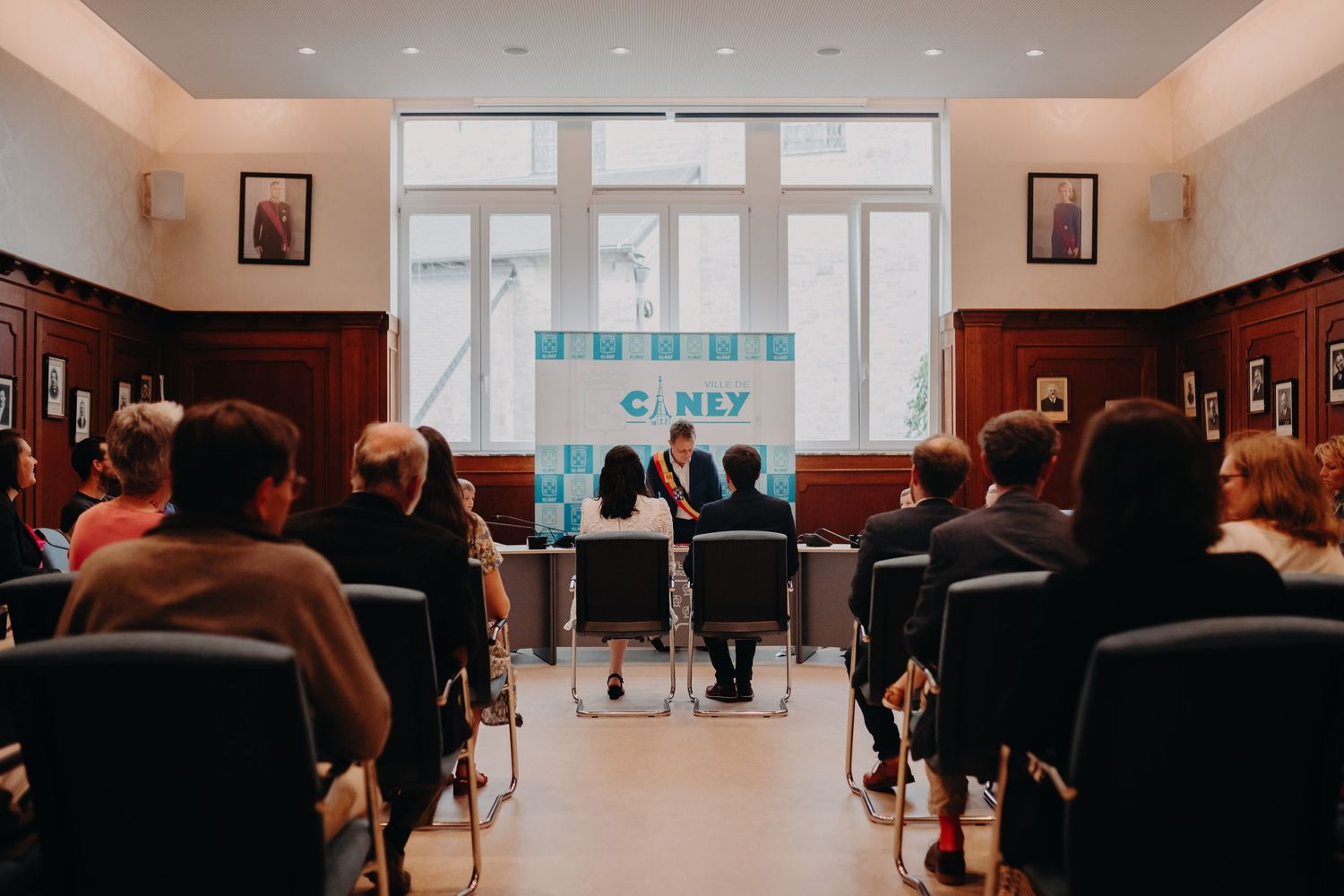 People gathered in a meeting room with red chairs and framed artwork on walls during what appears to be a presentation.