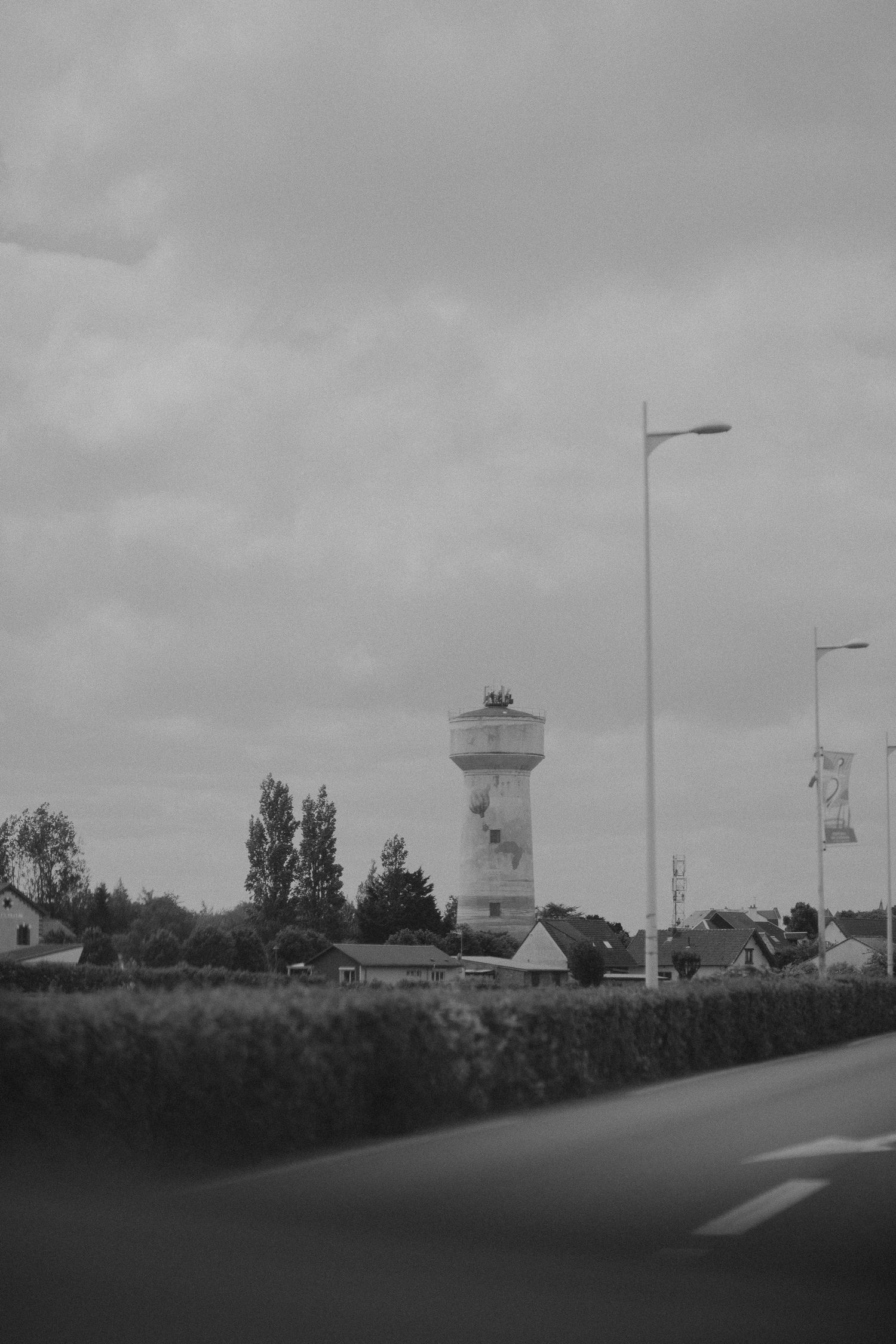 Black and white urban landscape with water tower and street lamps along a road under cloudy skies.